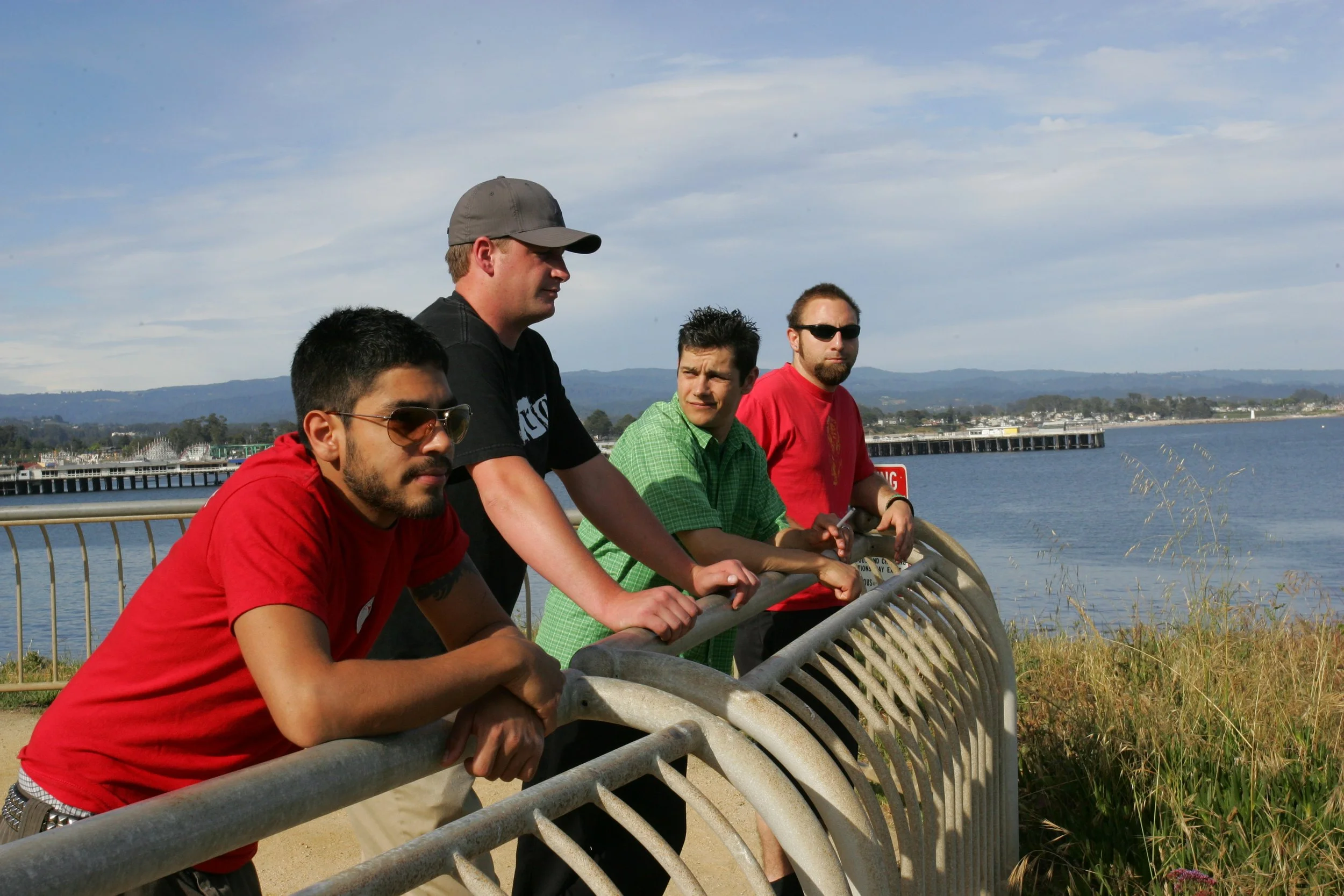 Four men standing by a fence overlooking a body of water with a pier and distant land in the background.