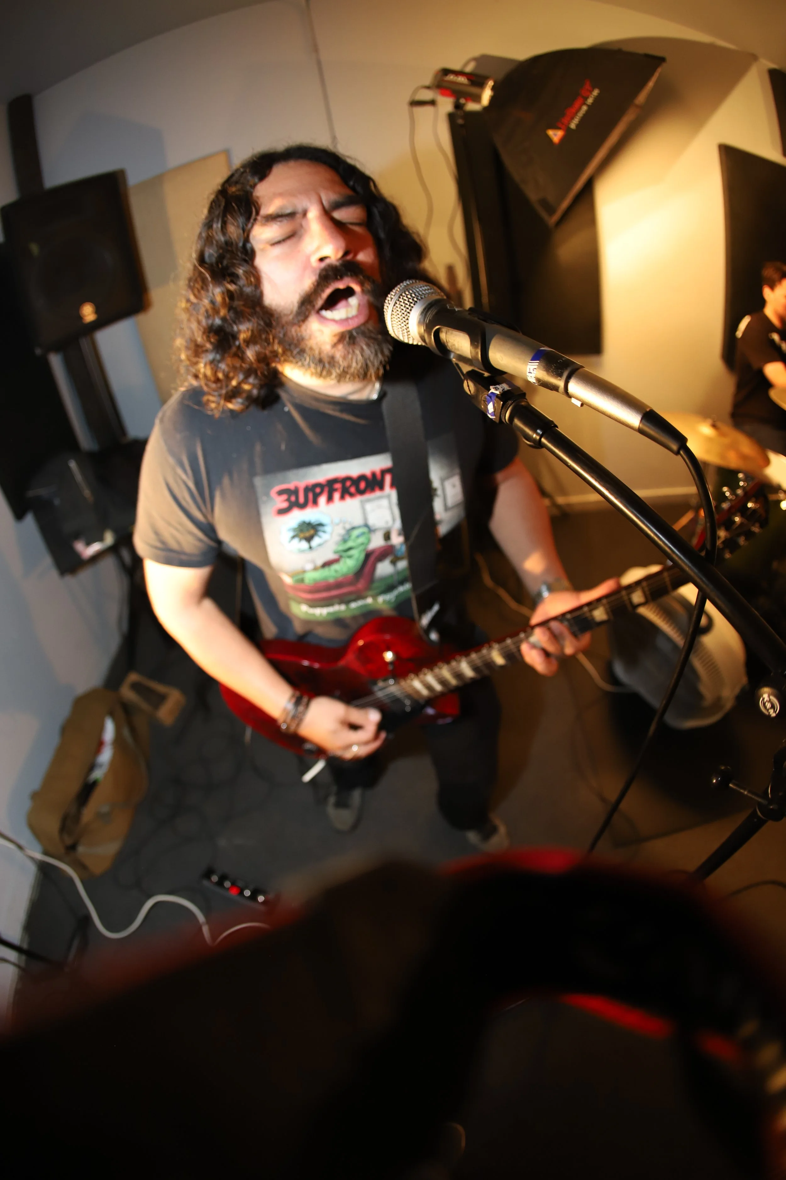 D$$ passionately singing into a microphone while playing an electric guitar. He is wearing a dark t-shirt and standing in a small, enclosed space with sound equipment and speakers around him.