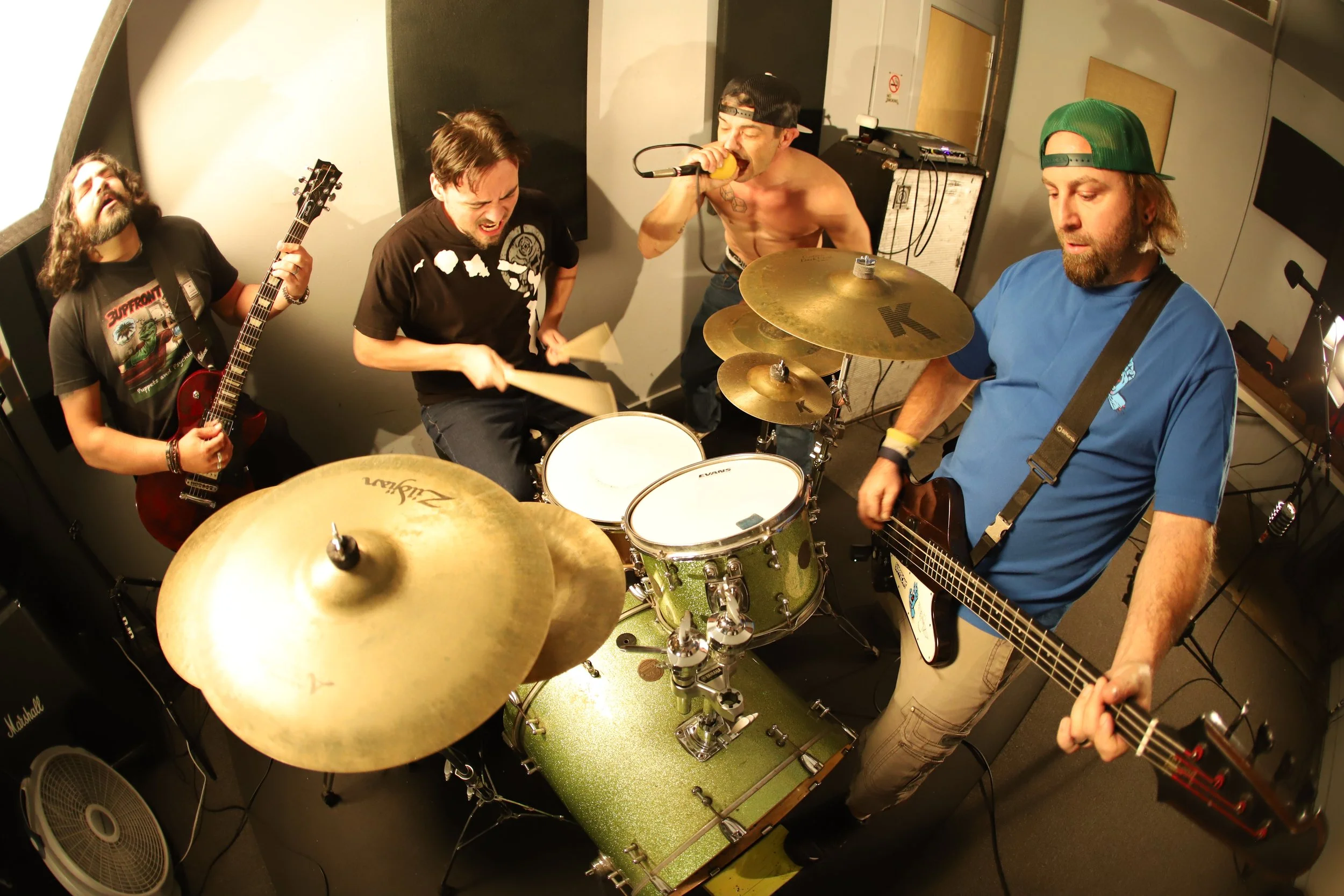 A band of five members performing in a small room. One person is playing guitar, another on drums, a third singing into a microphone, and the other two playing bass guitars. The room appears to be a practice space with soundproofing and musical equip