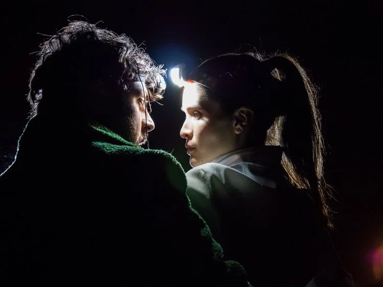 Close-up of a man and woman looking at each other with a headlamp on the man's forehead, in a dark setting