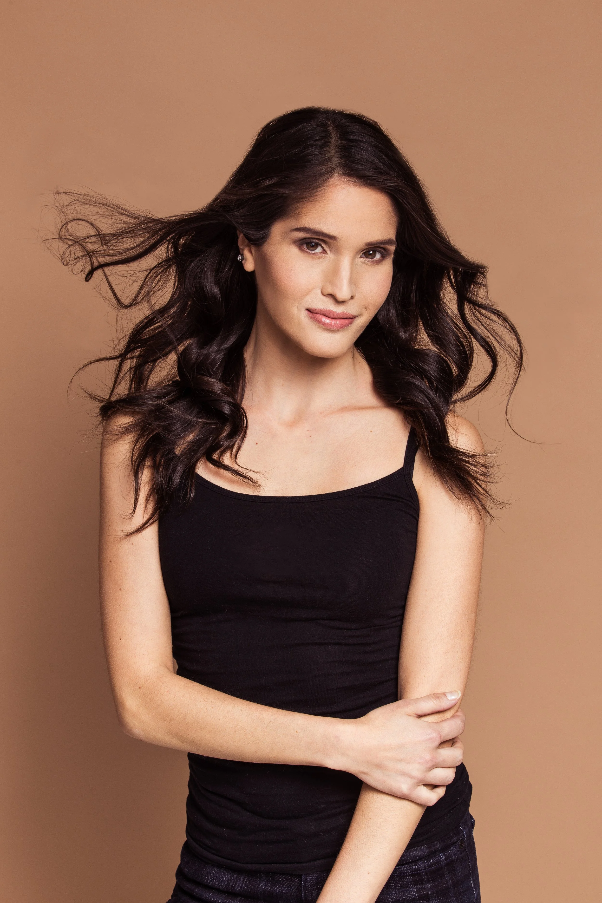 A woman with long, dark, curly hair wearing a black tank top and dark pants, standing against a beige background.