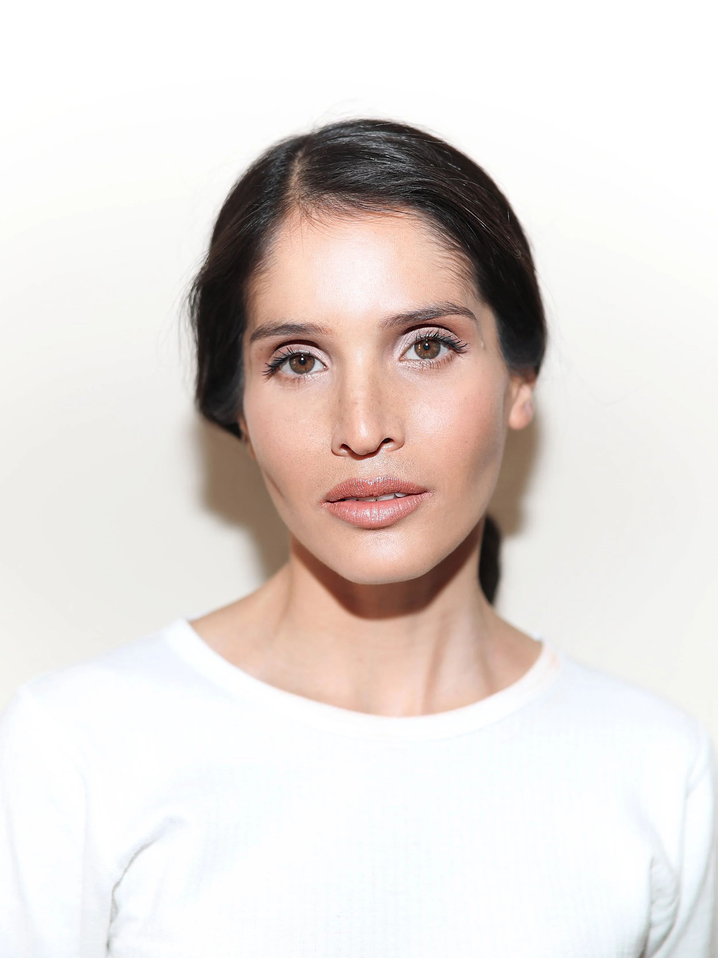 A woman with brown hair and brown eyes wearing a white top, standing against a plain white background.