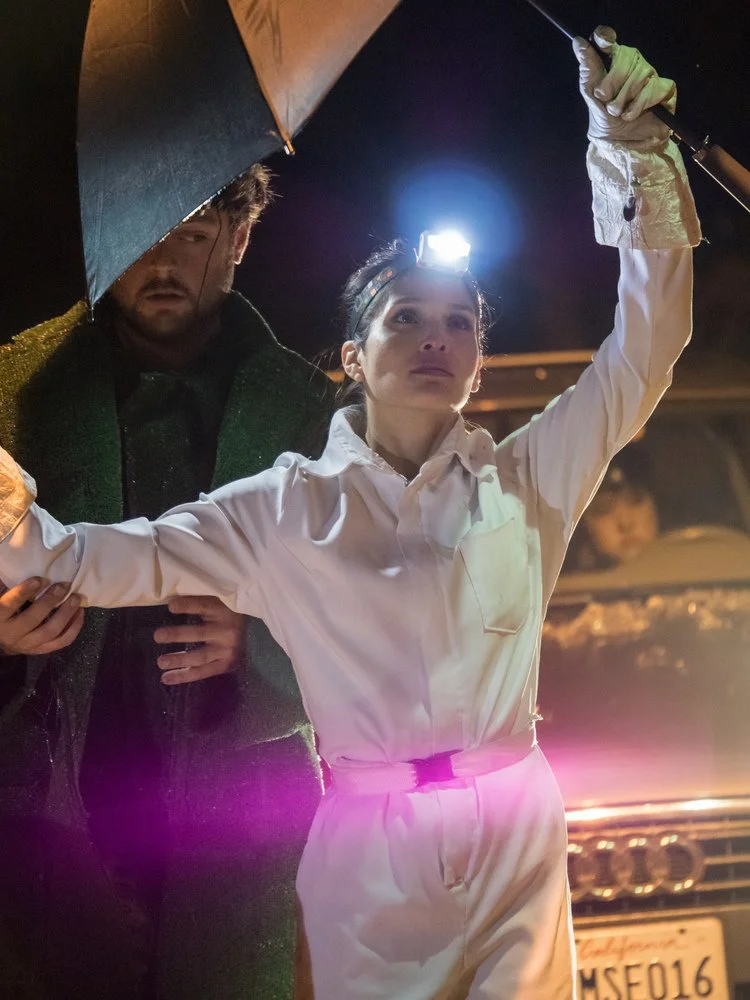 A woman in white scrubs and a headlamp holding a syringe, with an umbrella held by a man standing behind her at night.