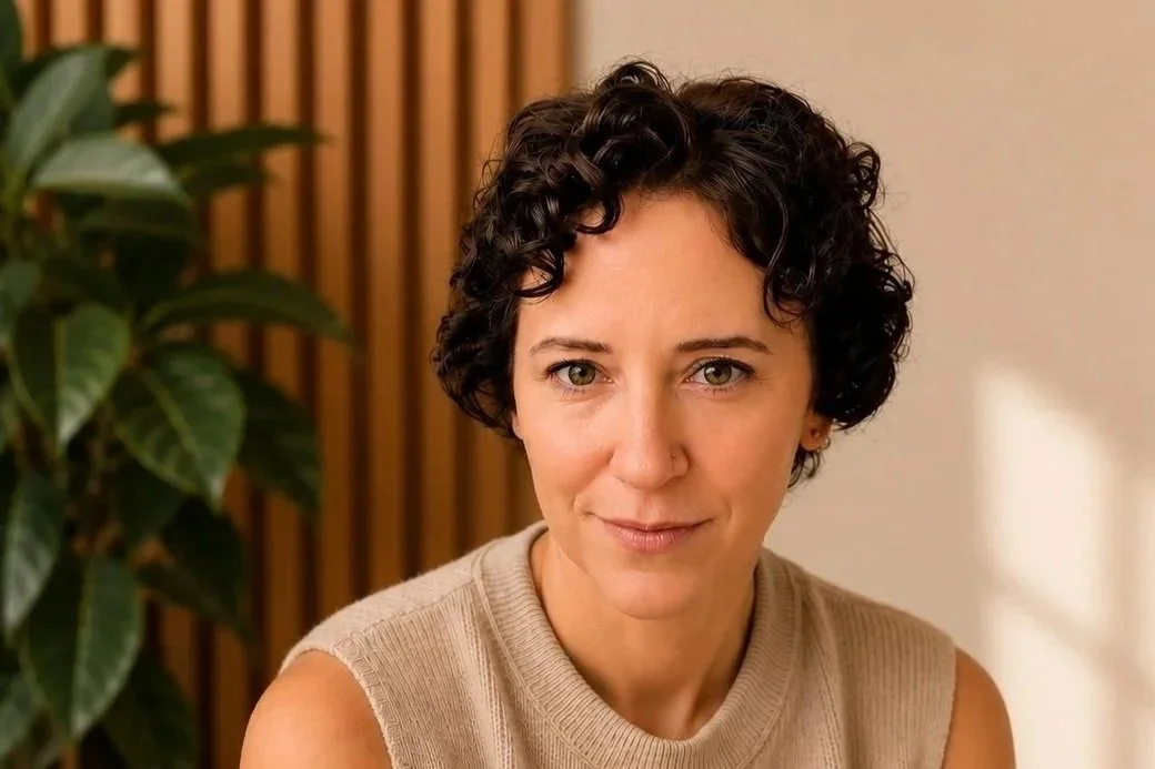 A woman with short, curly black hair, wearing a beige sleeveless top, sitting indoors with a plant and wooden slats in the background.