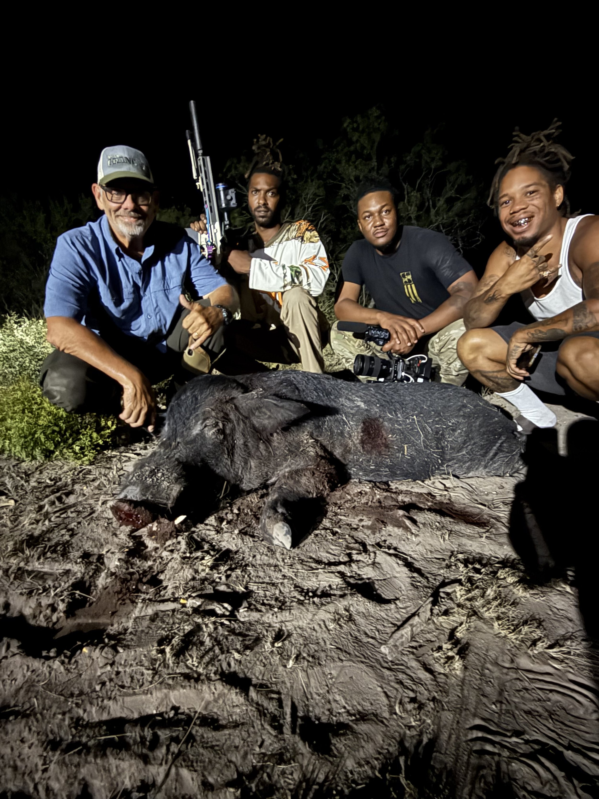 Four men kneeling outdoors at night around a dead pig on the ground, with trees and darkness in the background.