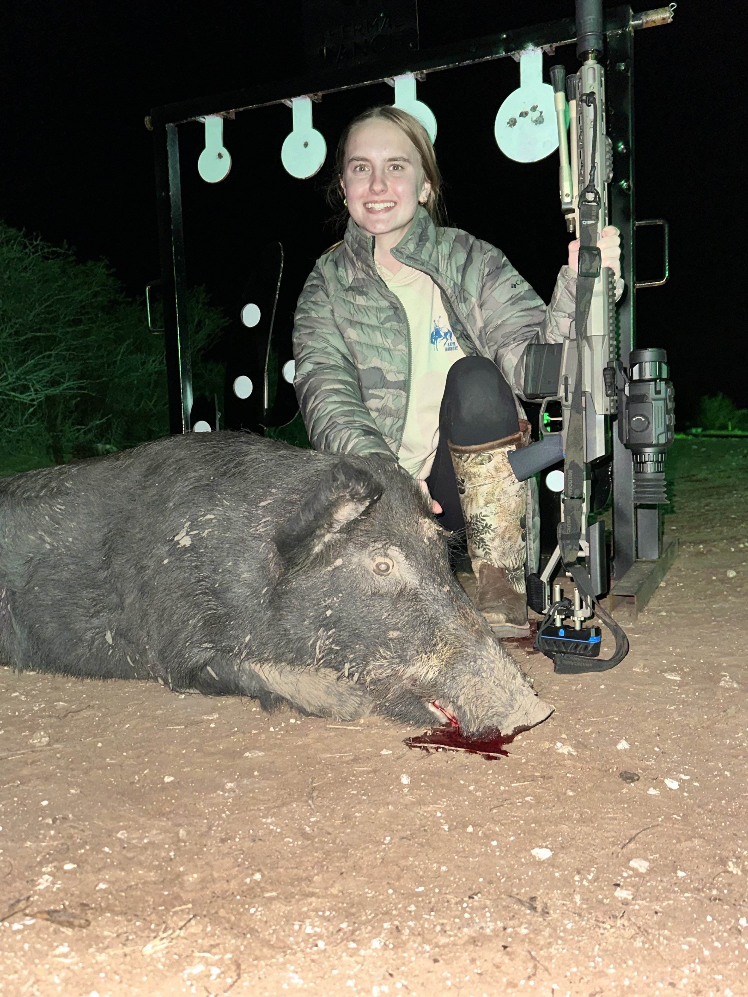 A woman is smiling and holding a rifle while kneeling next to a dead wild pig on the ground. The scene appears to be outdoors at night, with a shooting target stand and greenery in the background.