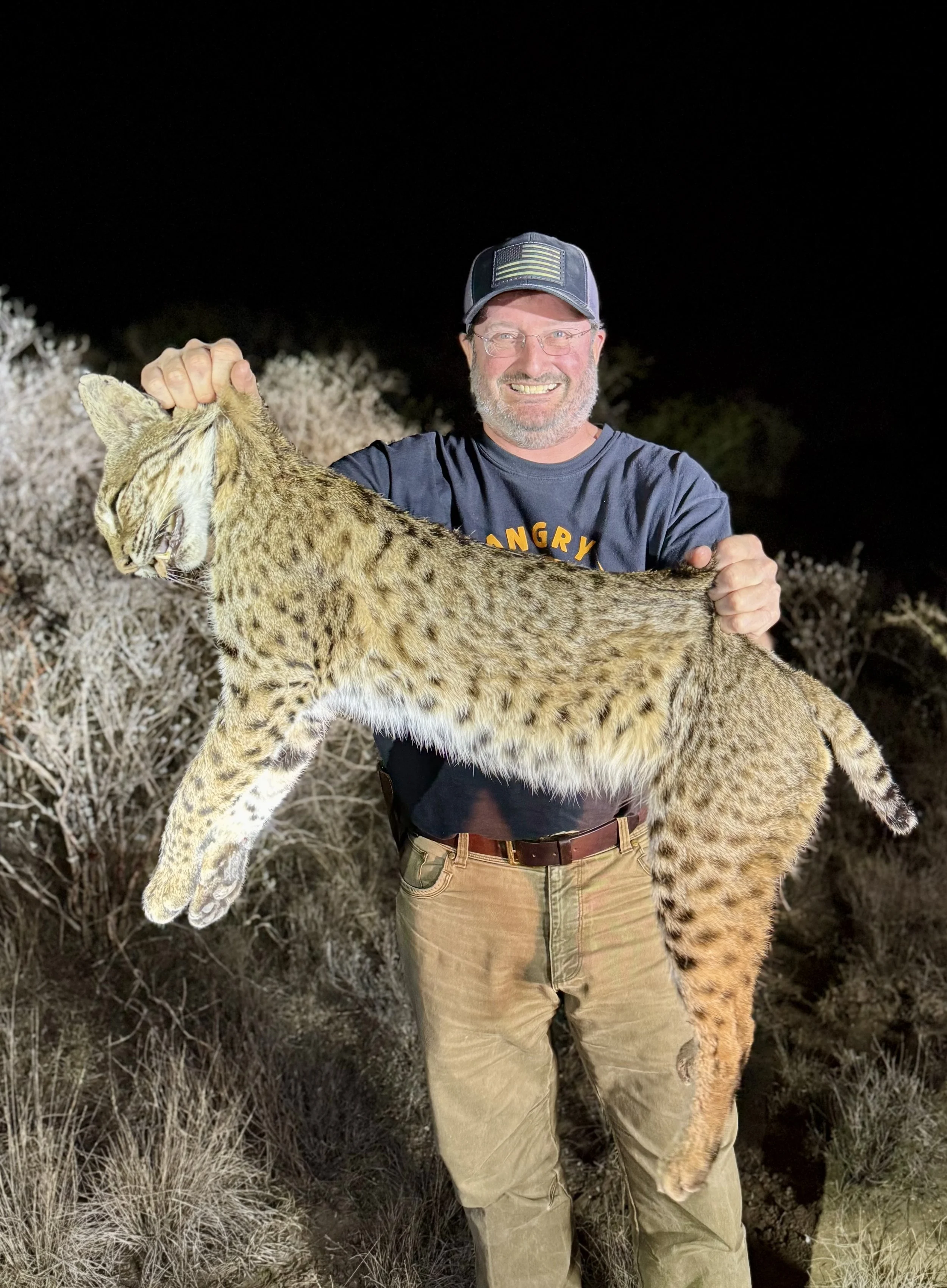 A man holding a dead mountain lion at night in a desert area, smiling and wearing glasses, a baseball cap, a dark shirt with yellow text, and khaki pants.