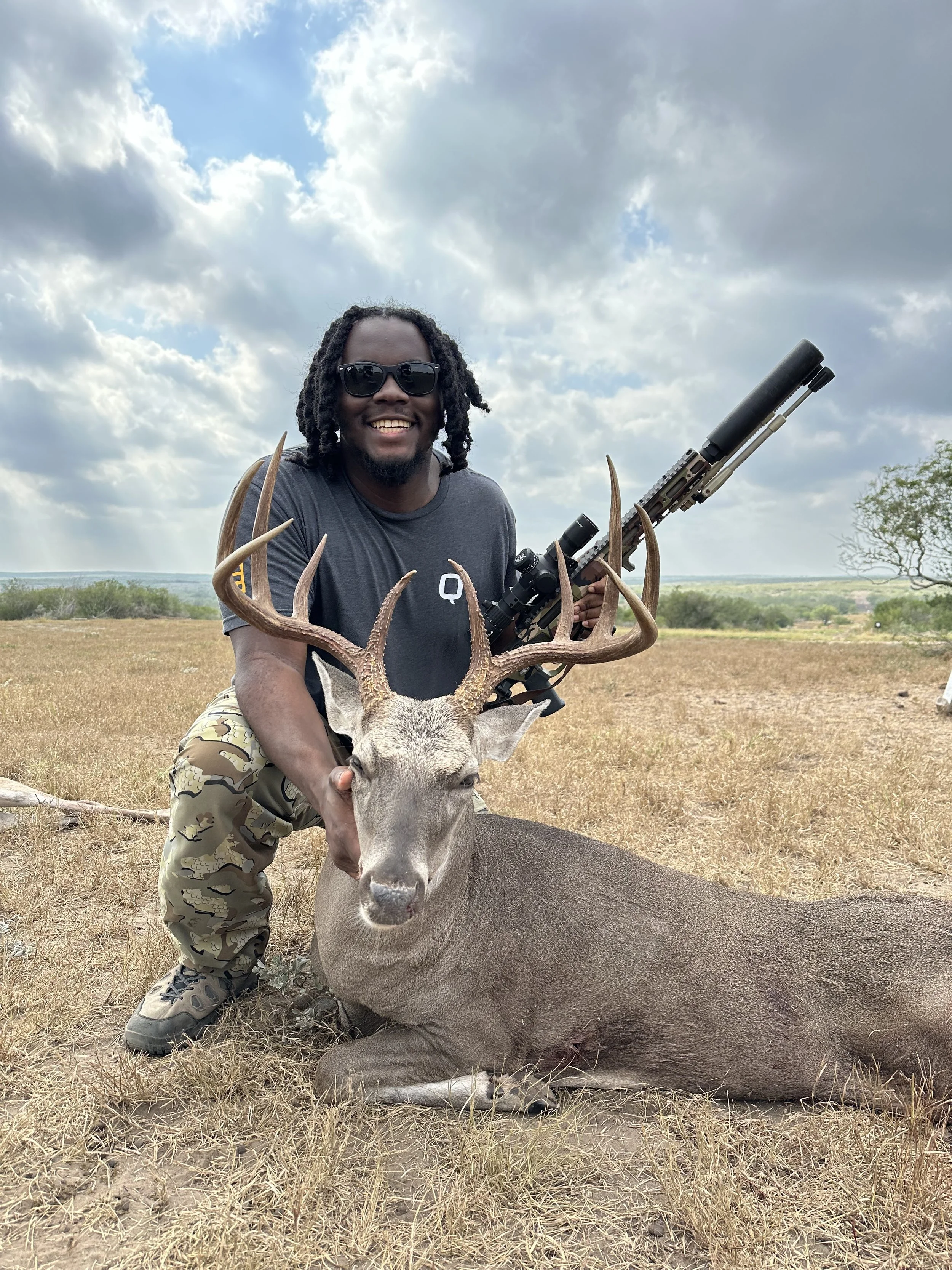 A man with dark skin, black curly hair, and sunglasses, smiling while kneeling next to a large deer with antlers in a dry, open field under a partly cloudy sky, holding the deer's antlers.