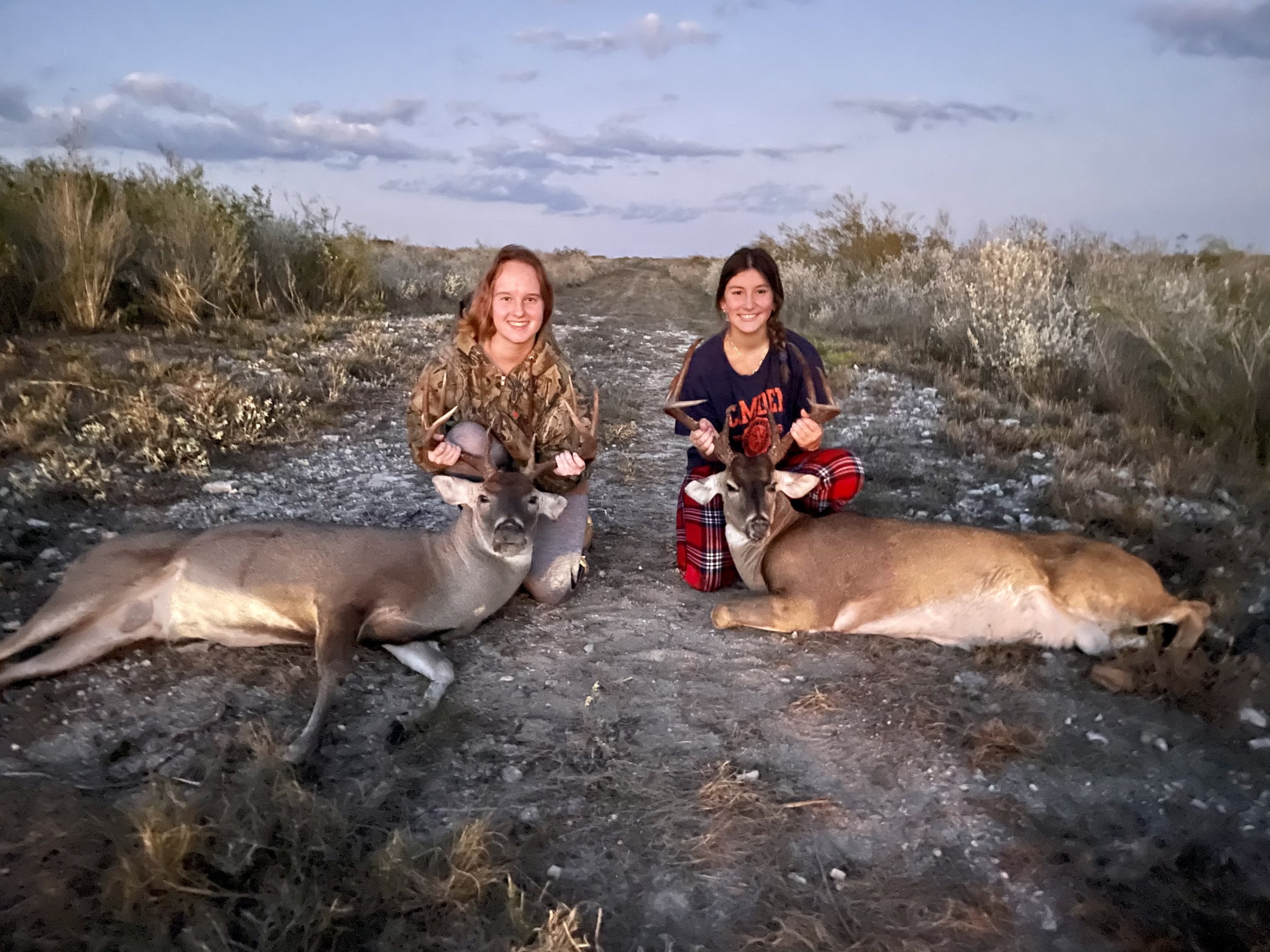 Two young women smiling and kneeling on the ground in a desert landscape during sunset, each holding a deer head mounted on a stick.