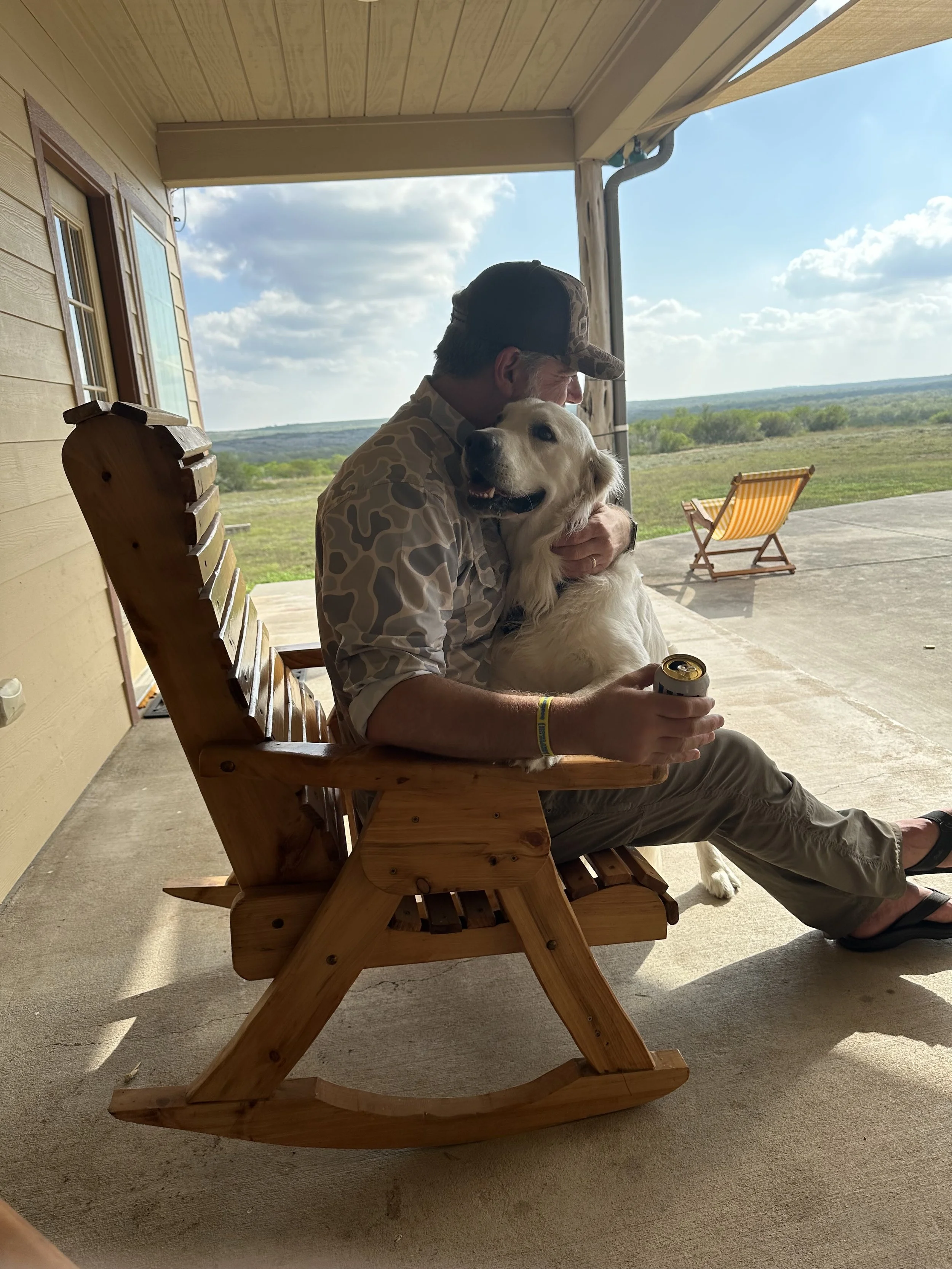 A man sitting on a wooden rocking chair on a porch, hugging a golden retriever dog. The man is wearing a camouflage cap and shirt, beige pants, and is holding a soda can. The background shows open landscape with green fields, a partly cloudy sky, and a yellow striped beach chair on the patio.