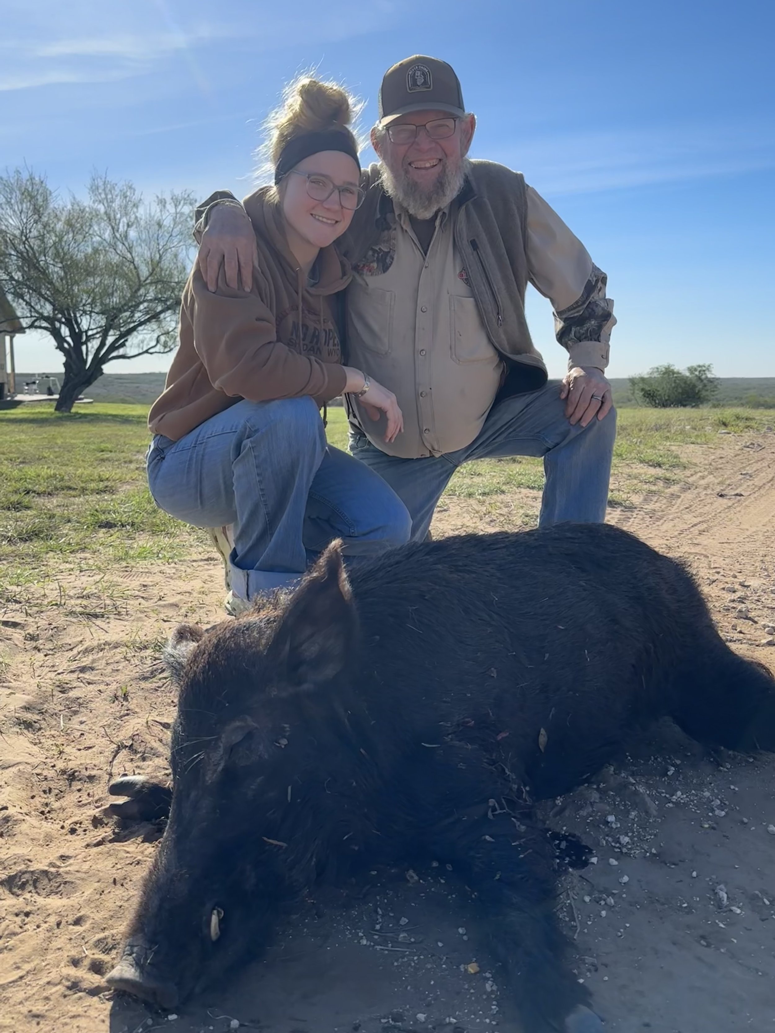 Two people, a young woman and an older man, kneeling on the ground outdoors, smiling at the camera with their arms around each other. There is a large black pig in the foreground, lying on the dirt. The background shows a clear blue sky, some trees, and a grassy area.