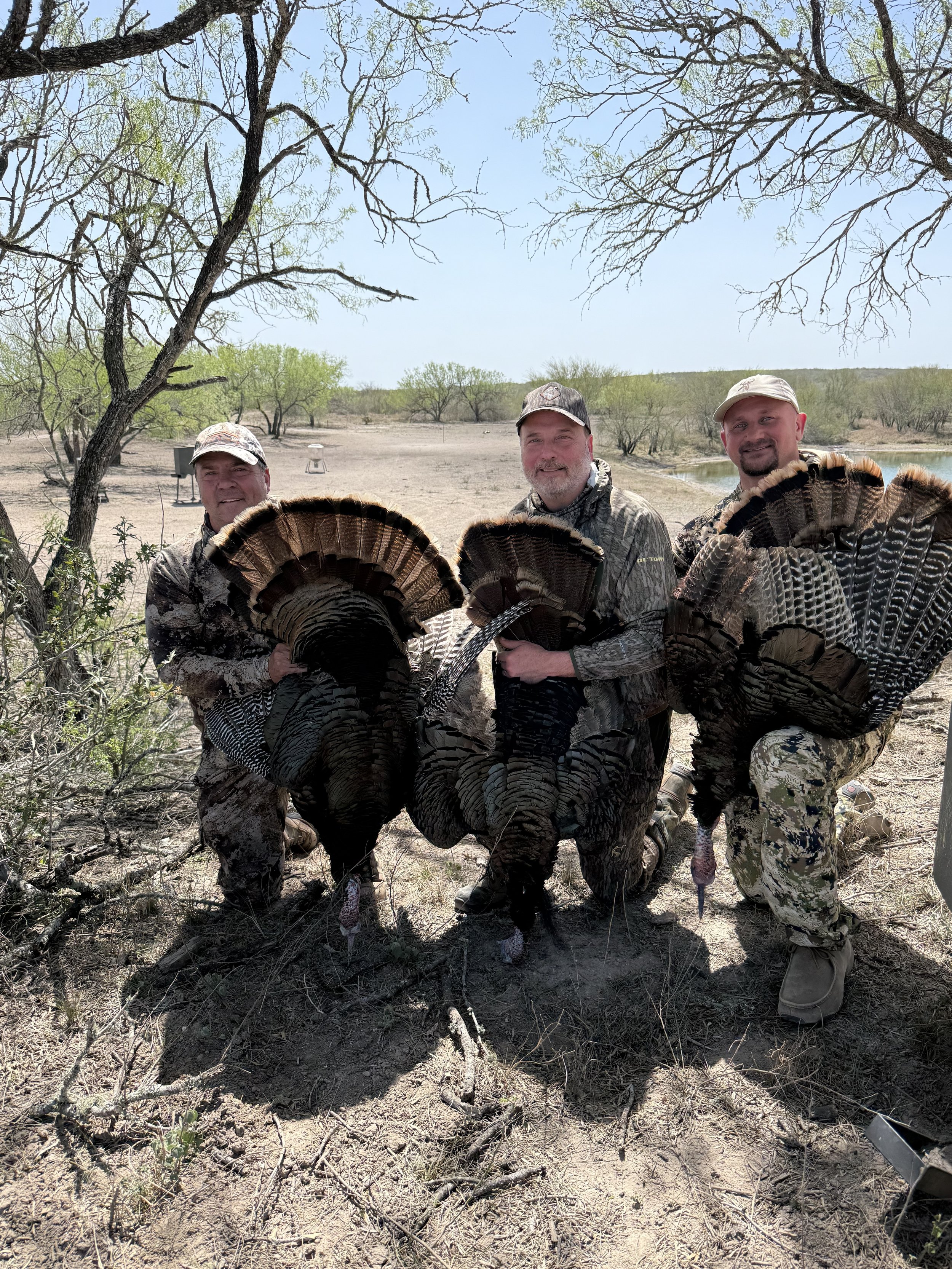 Three men in camouflage hunting clothes kneeling outdoors under trees, holding turkey fans and displaying a turkey they hunted, with open landscape, trees, and a small body of water in the background.