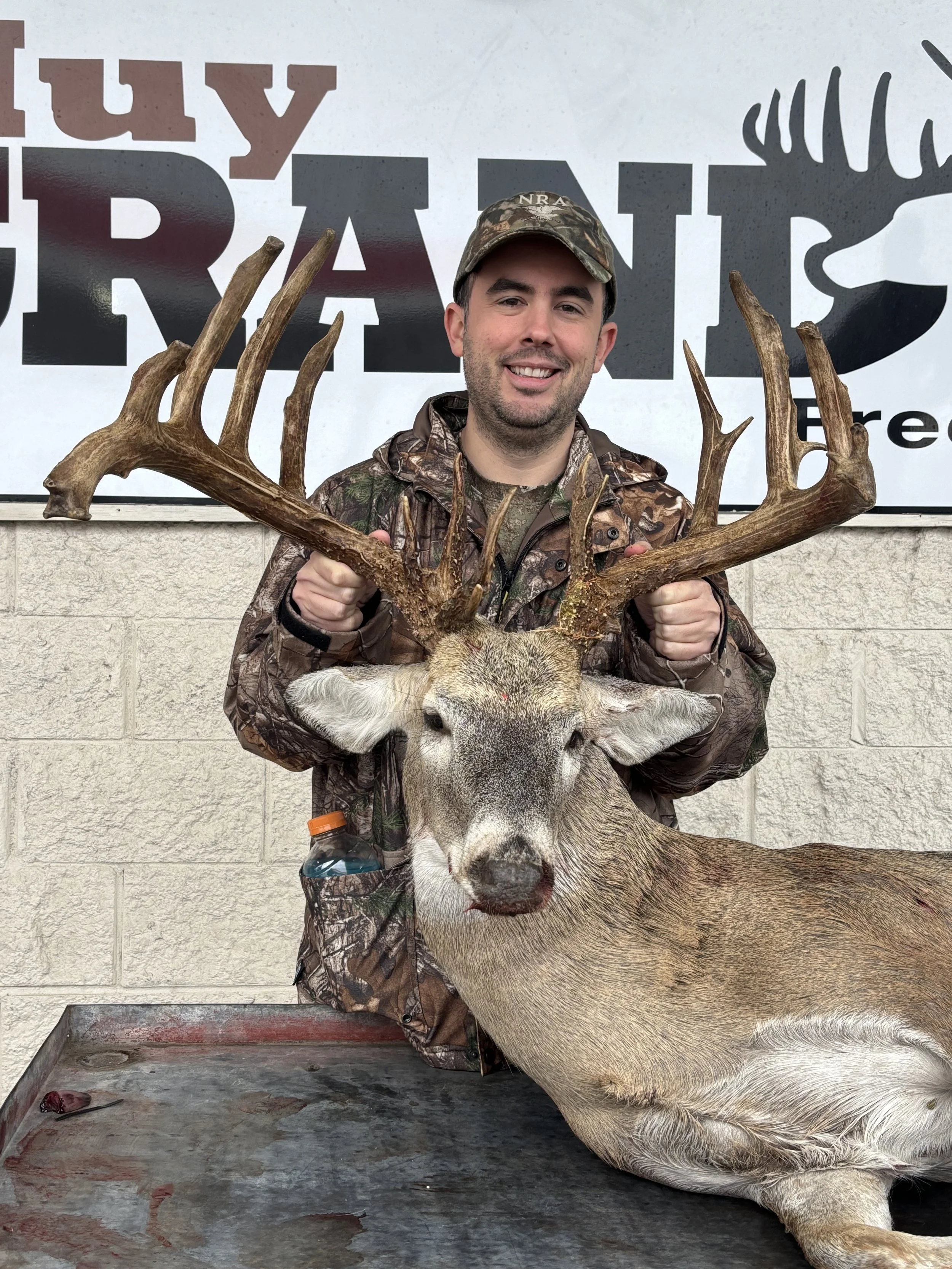 A man dressed in camouflage hunting gear is holding a large deer with antlers. The deer is lying on a bloodstained metal table. The man is smiling and standing in front of a sign that reads 'GUNNY's' and 'NRA' with a wolf silhouette.