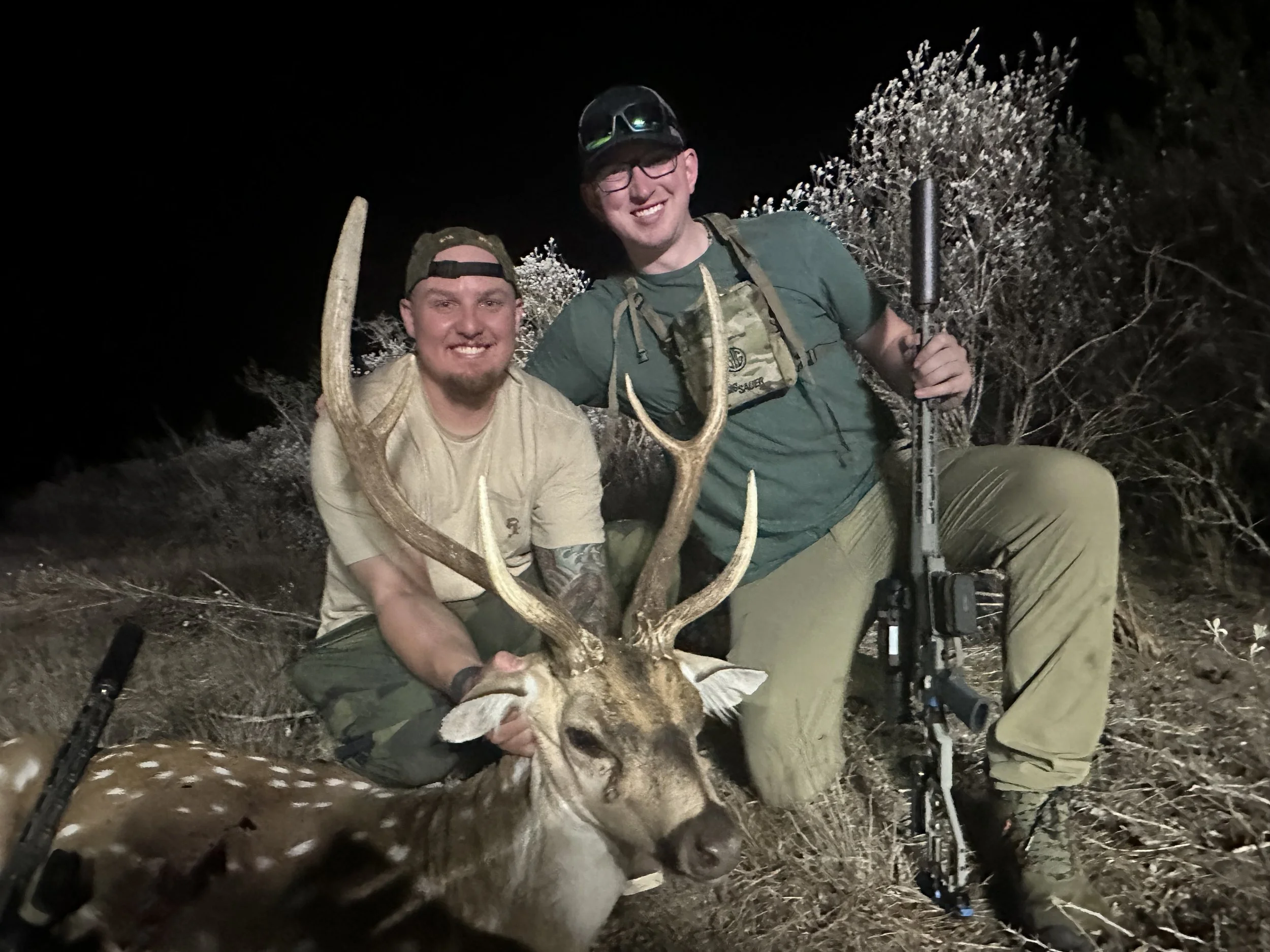 Two men smiling at night, one holding antlers and the other holding a rifle, with a fox in front of them and bushes in the background.