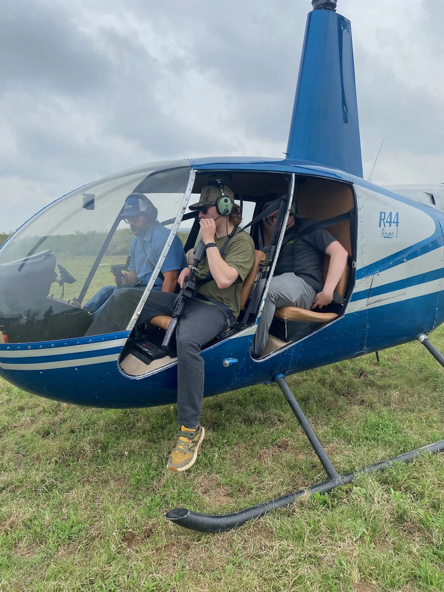 Three men inside a blue and white helicopter on a grassy field, with one man wearing a green shirt and headphones, another in a blue shirt and cap, and the third in a black shirt, under a cloudy sky.