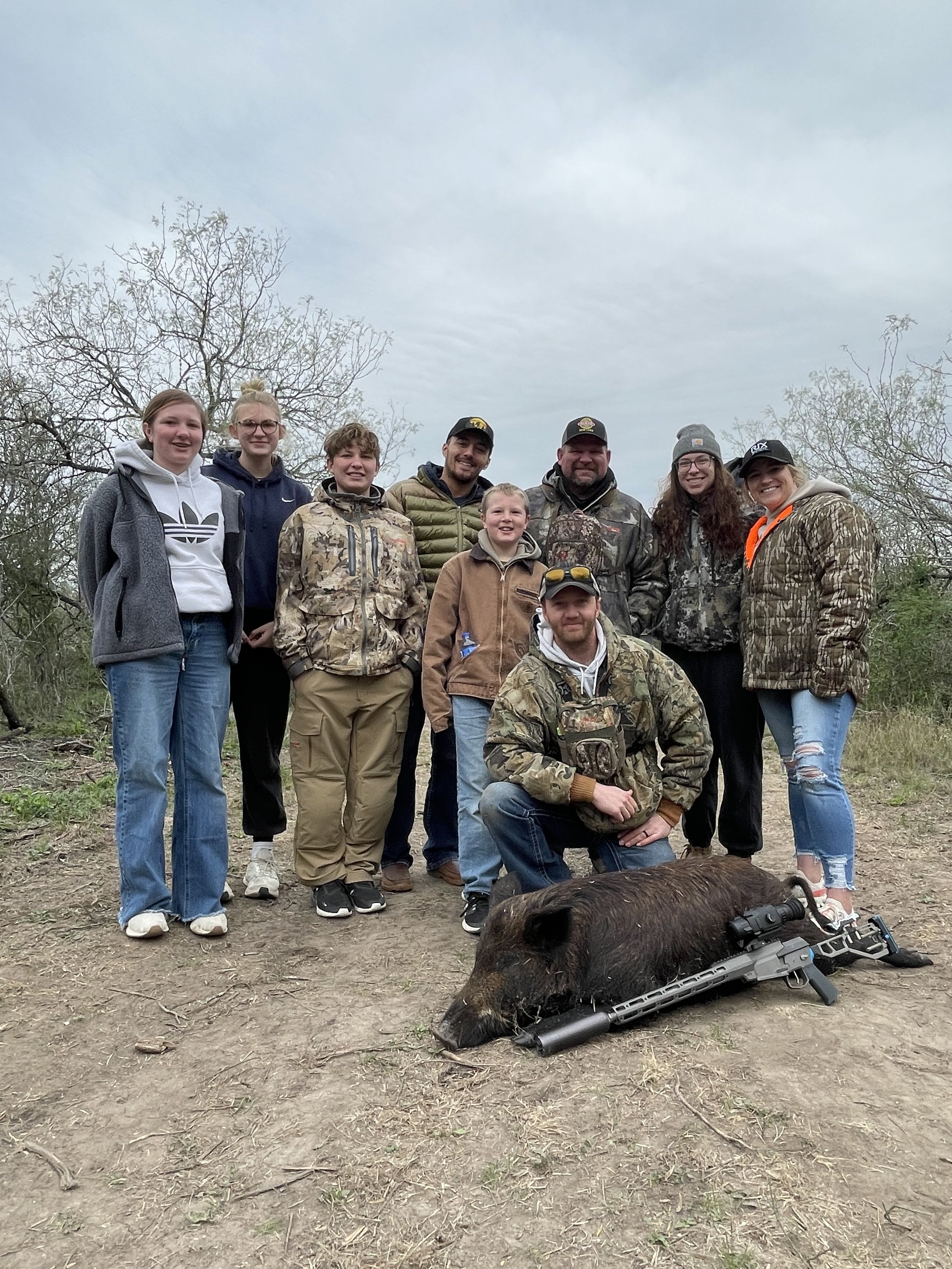 A group of eight people, including children and adults, standing outdoors in a natural setting with trees and cloudy sky, posing behind a large dead pig with a rifle in front of it.