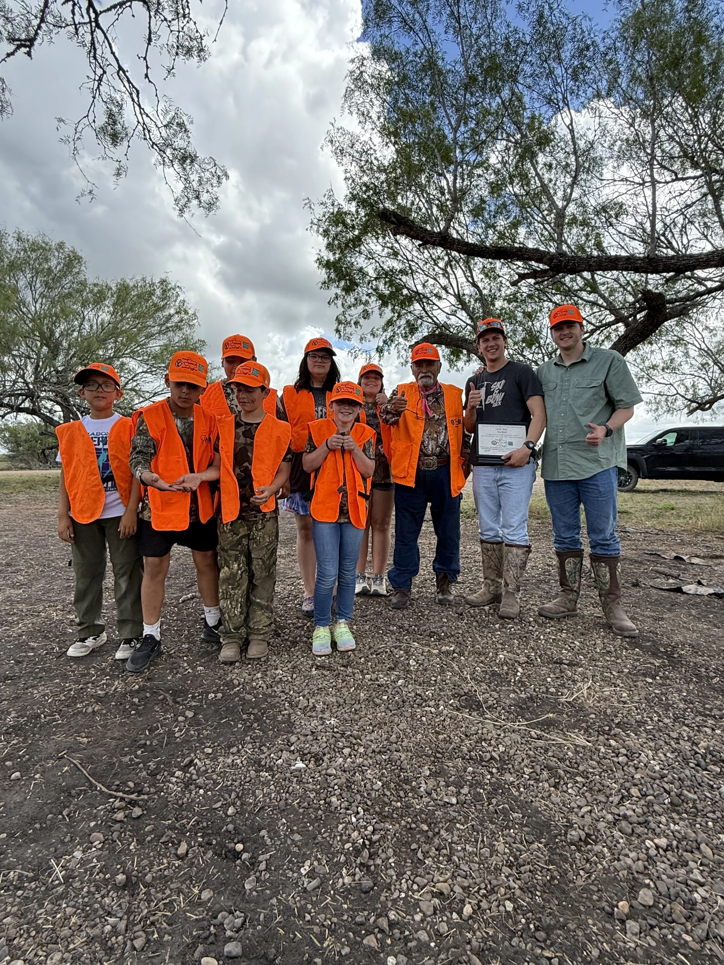 Group of ten people, including children and adults, standing outdoors on a rocky ground under a partly cloudy sky with trees in the background. They are wearing bright orange safety vests and caps, and some are holding tools or certificates, indicating they may be participating in an outdoor or conservation activity.