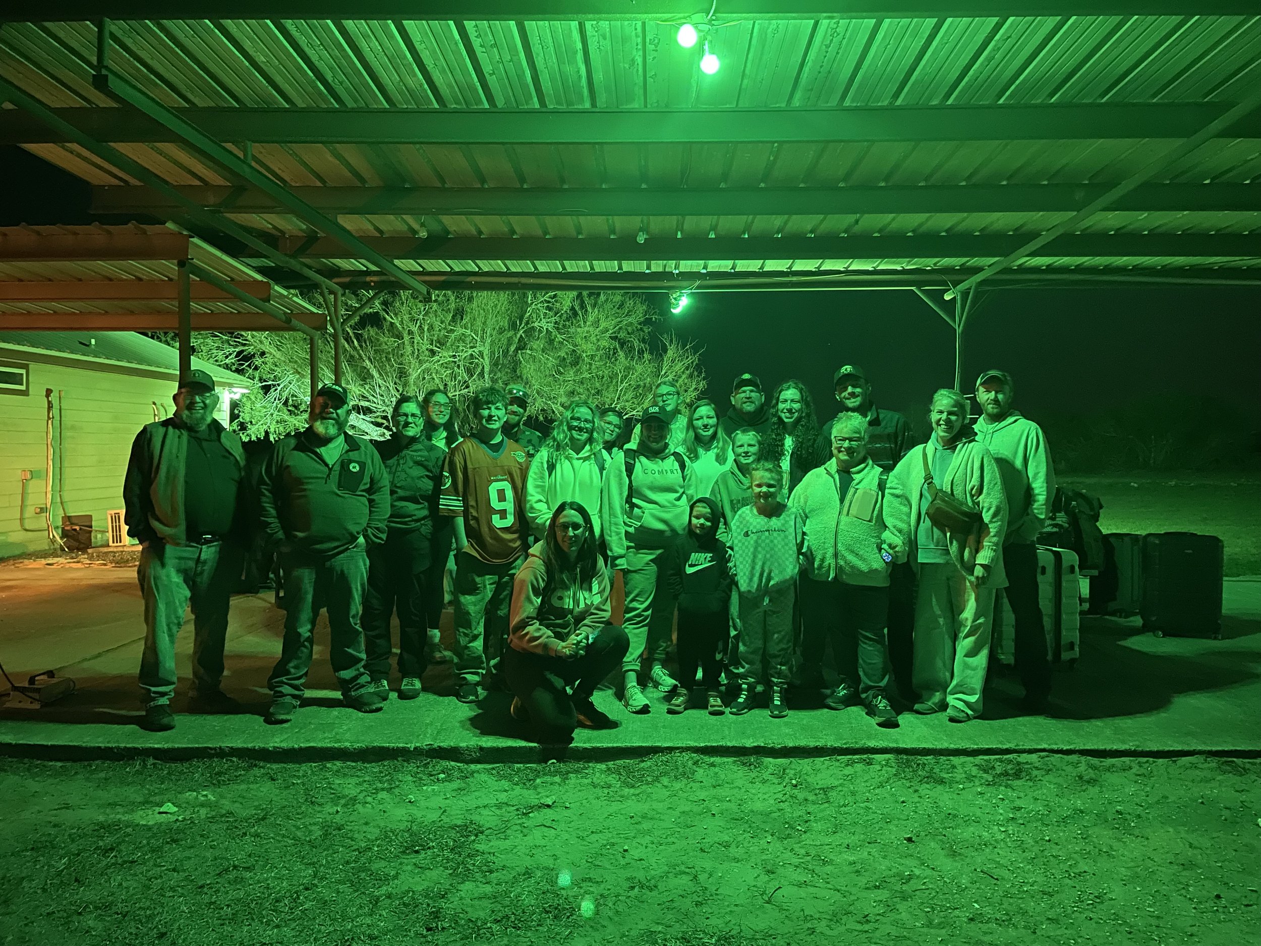Group of people standing outdoors at night under a metal roof, illuminated by green lighting, with trees in the background.