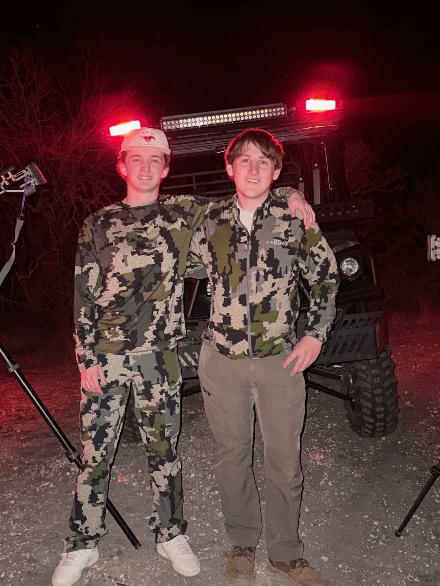 Two young men standing outdoors at night, dressed in camouflage military-style clothing, with a vehicle and equipment behind them, illuminated by red emergency lights.