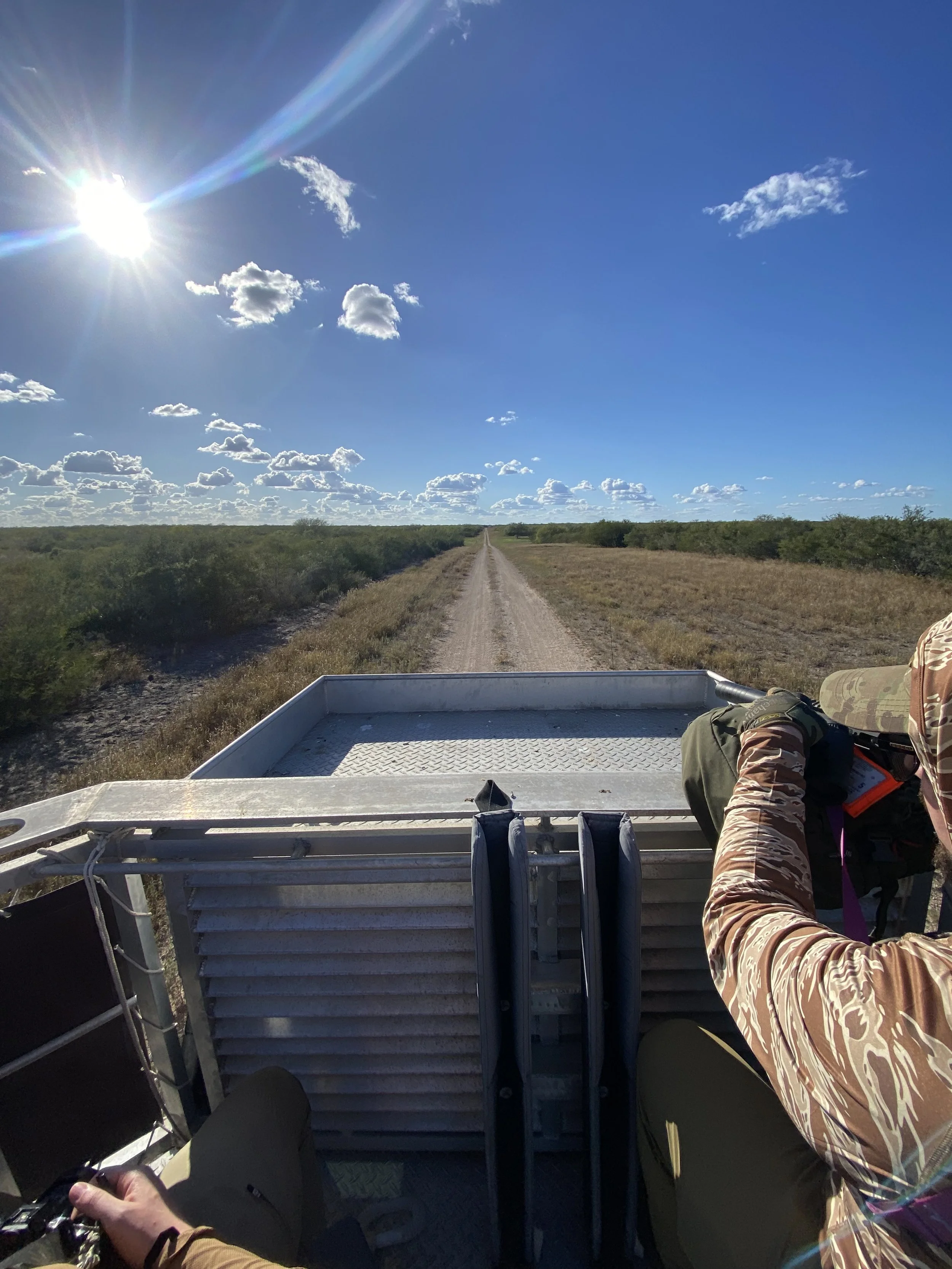 The photo shows the front of a wildlife or park vehicle with navigation screen, the person driving is wearing camouflage clothing, and the vehicle is on a dirt road through open flat land with sparse bushes, under a bright sunny sky with some clouds.