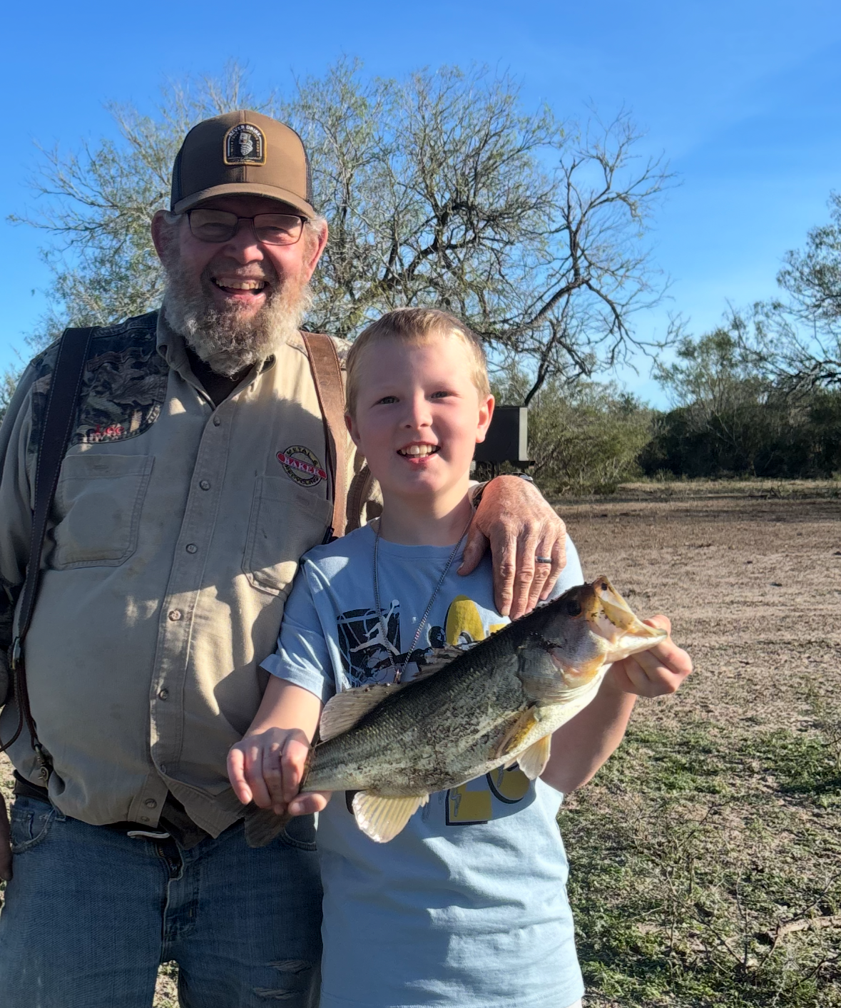 An older man and a young boy smiling outdoors, holding a large fish.