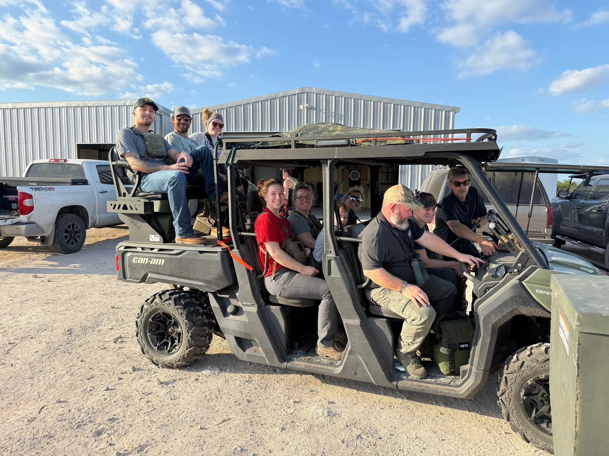 A group of people riding in two off-road utility vehicles parked in front of a metal building on a sunny day with blue sky and clouds.
