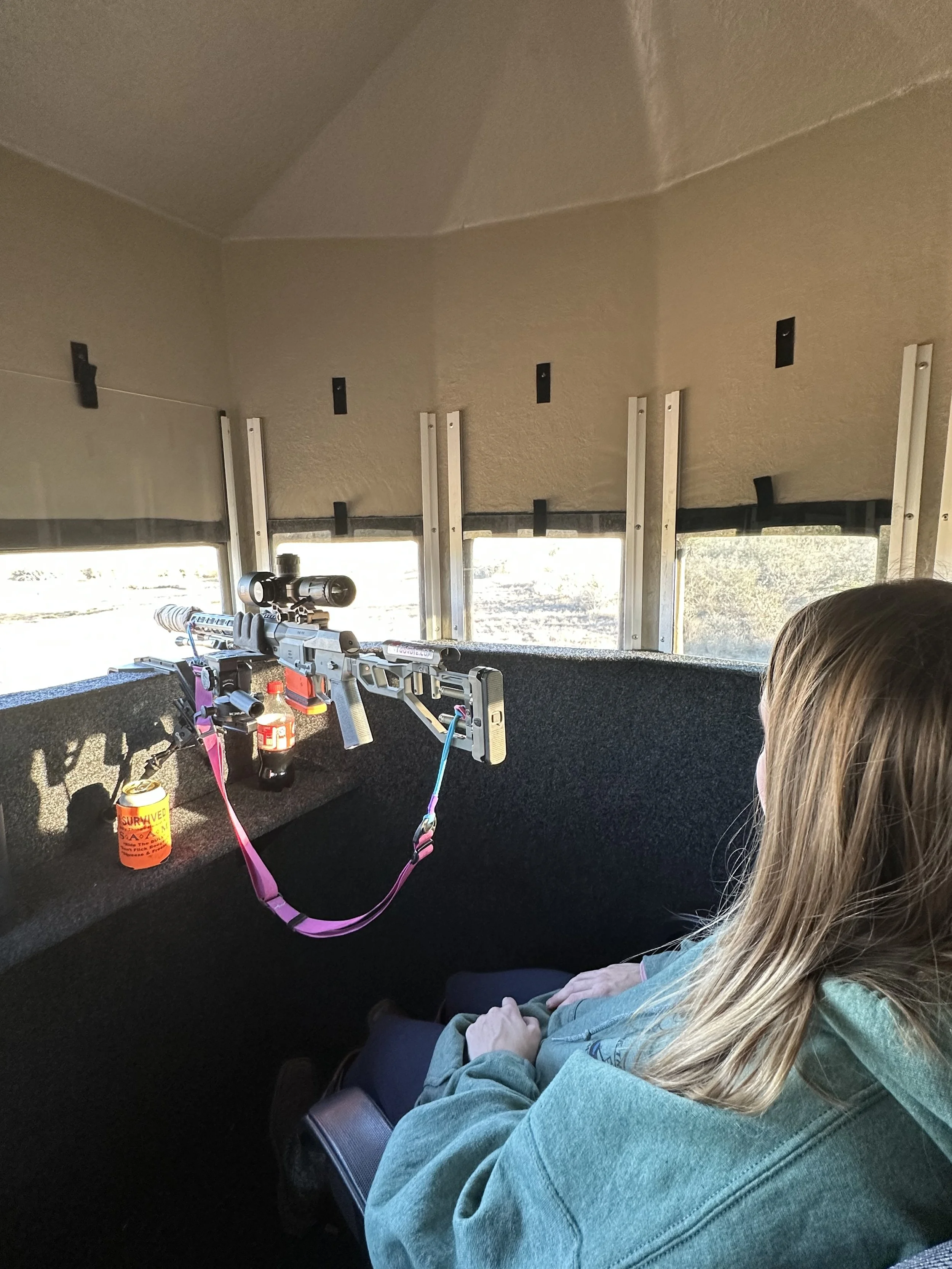 Inside a shooting range with a scoped rifle resting on a stand, a drink bottle, and a person's hair visible in the foreground, and windows with a landscape view outside.