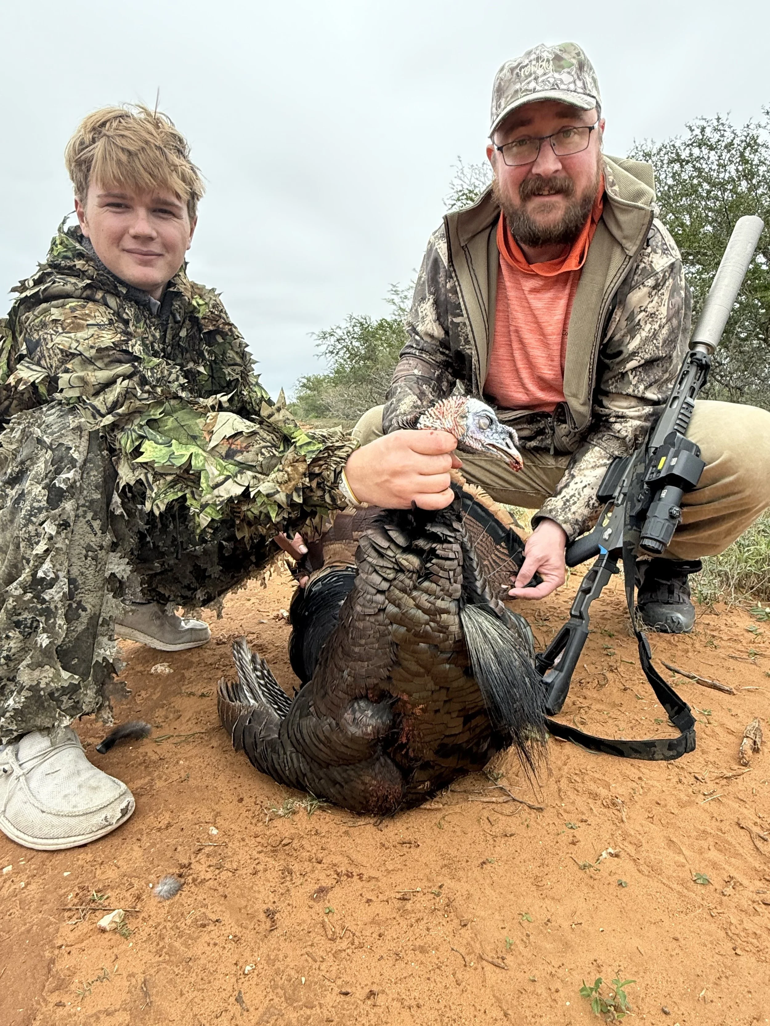 Two men posing with a large wild turkey on sandy ground, surrounded by desert vegetation, during daytime with an overcast sky.