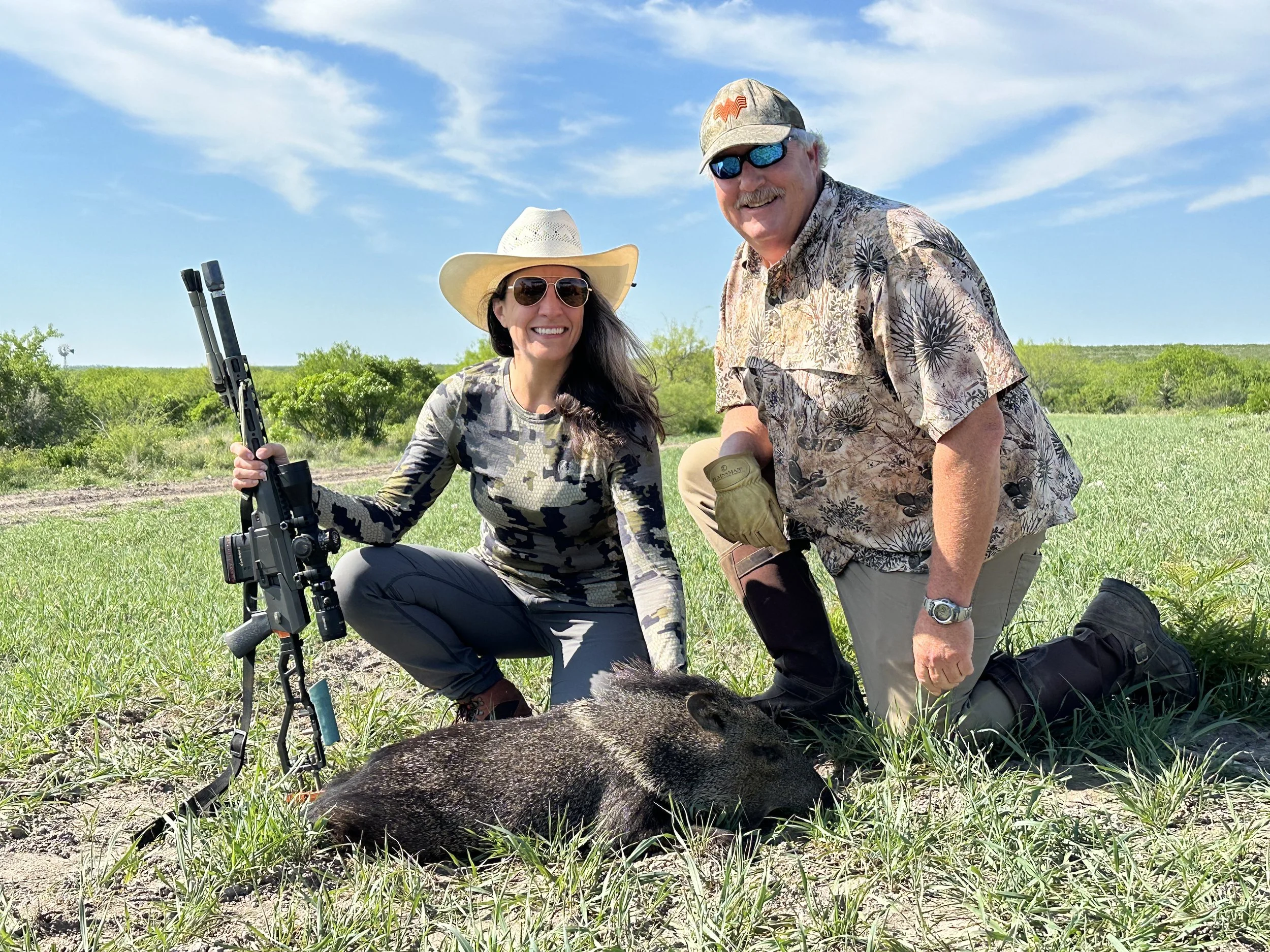 Two people outdoors in a field, holding rifles, wearing camouflage clothing, hats, and sunglasses, posing with a dead boar on the ground.