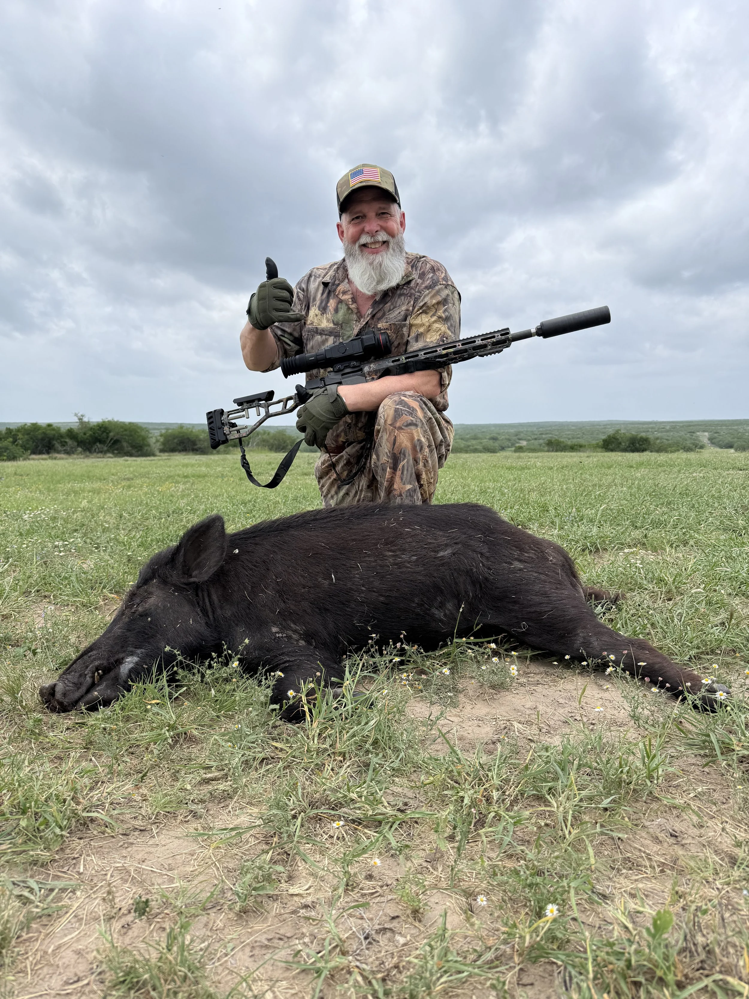 A man dressed in camouflage clothing with a gray beard kneeling on the grass, holding a rifle, and giving a thumbs-up while smiling. There is a dead pig lying on the ground in front of him. The background features a cloudy sky and a rural landscape.