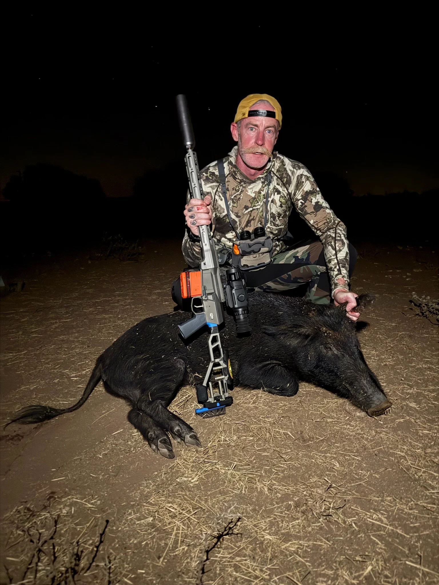 A man with a yellow cap, camouflage clothing, and binoculars around his neck kneels on the ground at night, holding a rifle, with a large black pig lying on the dirt.