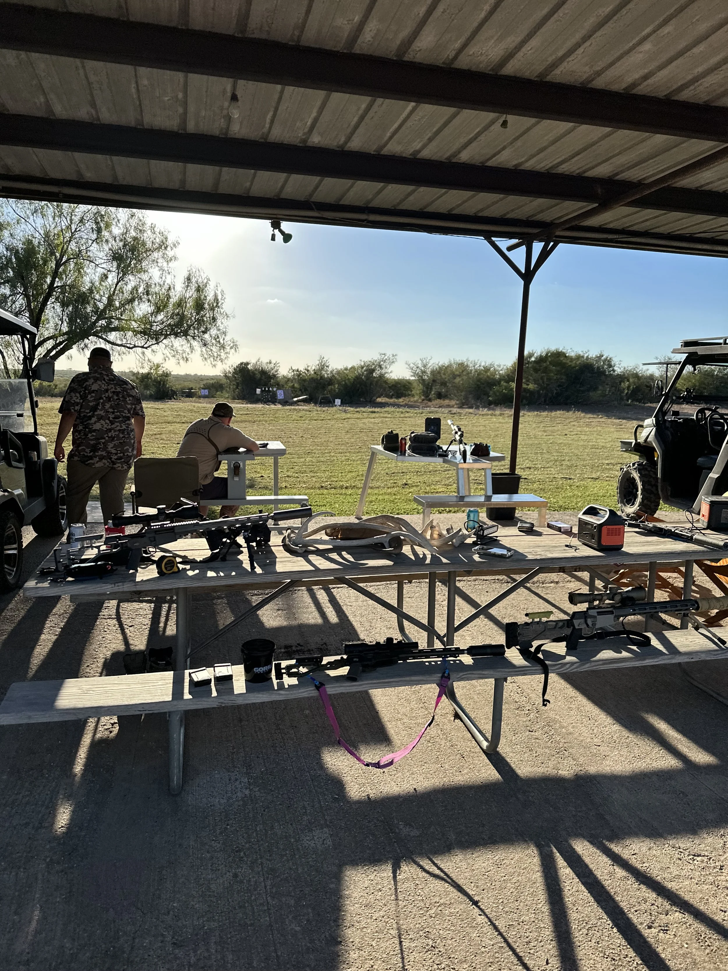 Outdoor shooting range with targets set up in the distance, covered by a metal roof, and tables in the foreground displaying firearms, ammunition, and shooting accessories.