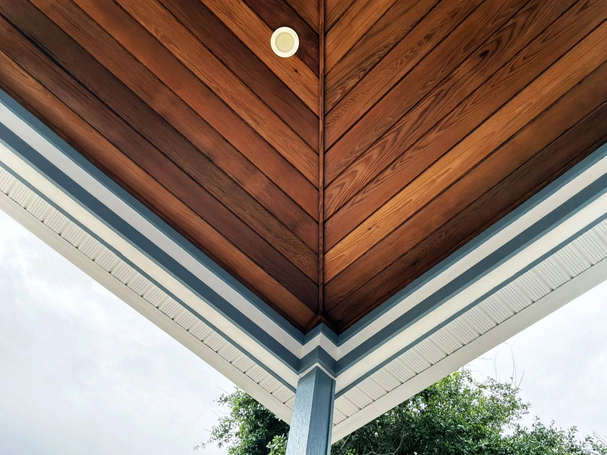 View of the underside of a wooden porch roof with a small round ceiling light fixture and decorative white trim, with trees visible in the background.