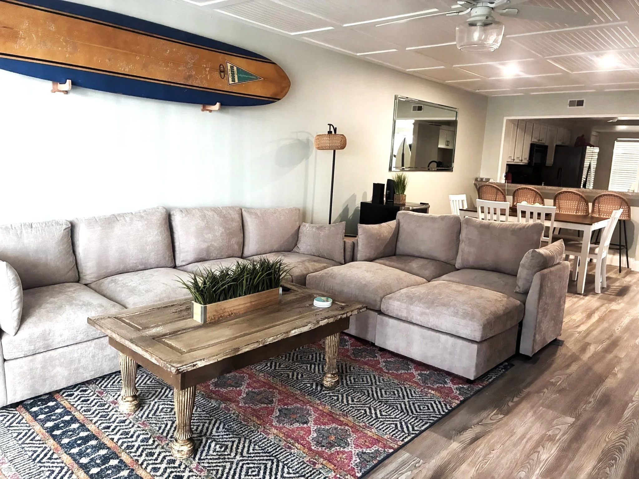 Living room with an L-shaped beige sofa, a rustic wooden coffee table with gold accents, a patterned area rug, a floor lamp, a wall mirror, and a fish-shaped wooden surfboard mounted on the wall. Visible in the background is an open kitchen with white cabinetry and a dining area with wicker chairs.