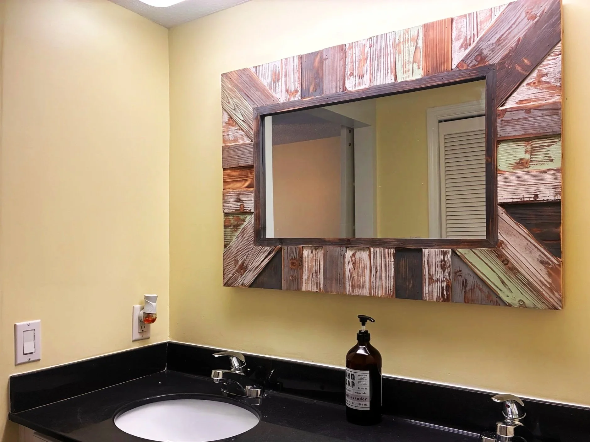 A bathroom vanity with a black countertop, a white sink, and two silver faucets. A brown soap dispenser bottle is on the right side of the counter. A mirror with a rustic wooden frame made of reclaimed wood planks is hung on a light yellow wall, with part of a white door with slats visible in the reflection.