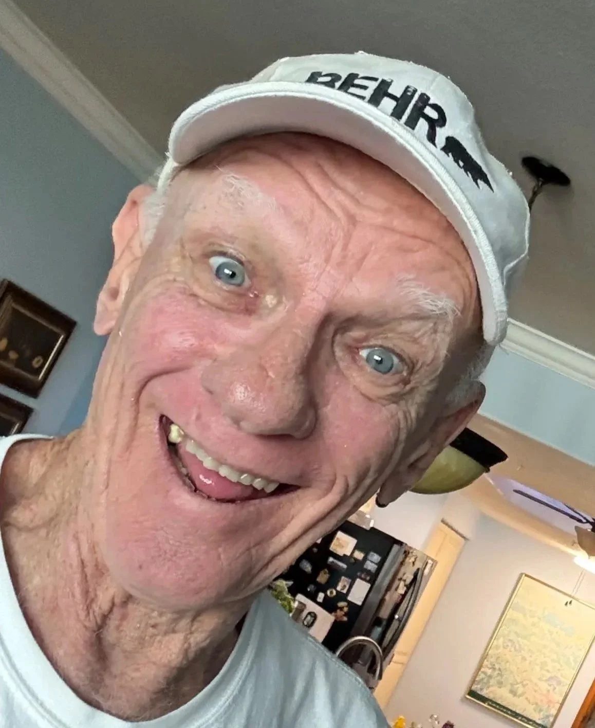 A close-up photo of an elderly man with blue eyes, white hair, and wearing a white cap with black text, smiling with a missing front tooth, in a home kitchen setting.