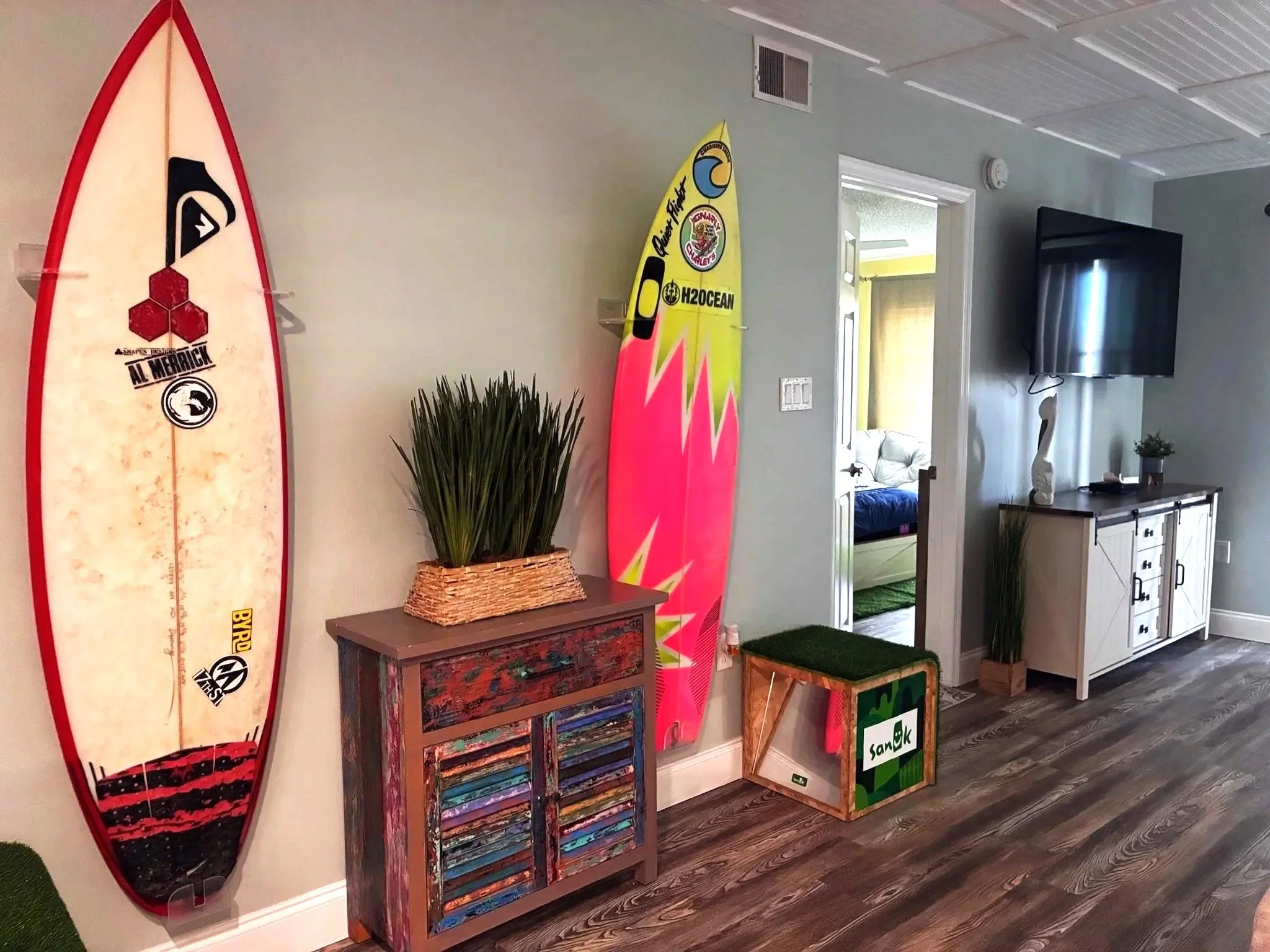 Indoor room corner with surfing-themed decor, including a surfboard with various logos, a colorful wooden cabinet, framed artwork, a white cabinet, and potted plants.