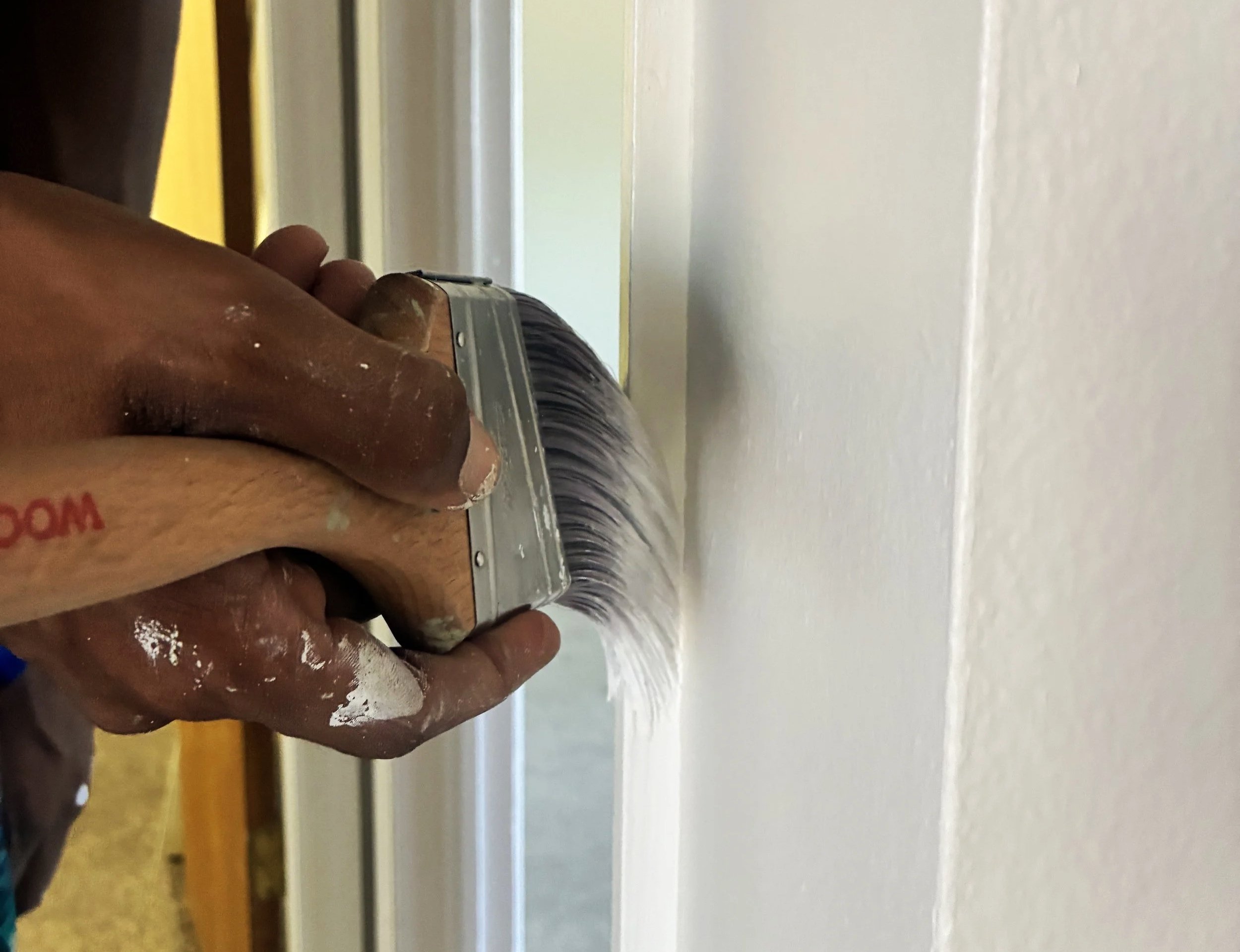 Person sanding a wall with a hand-held electric sander, visible hand with some paint or dust on the fingers.