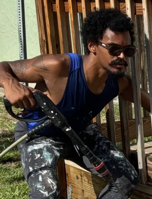 Man with curly hair, sunglasses, tattoos, wearing a blue sleeveless shirt and paint-splattered pants, working with a power tool on a wooden structure outdoors.