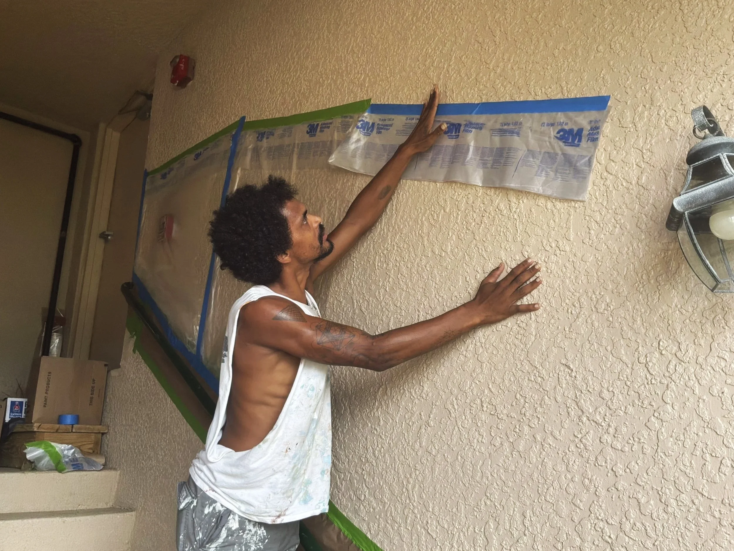 A man with curly hair and tattoos on his arms installs a decorative molding on a beige textured wall. He is wearing a white tank top and is reaching up with his left hand to work on the molding, which has packaging from 3M in blue and green tape.