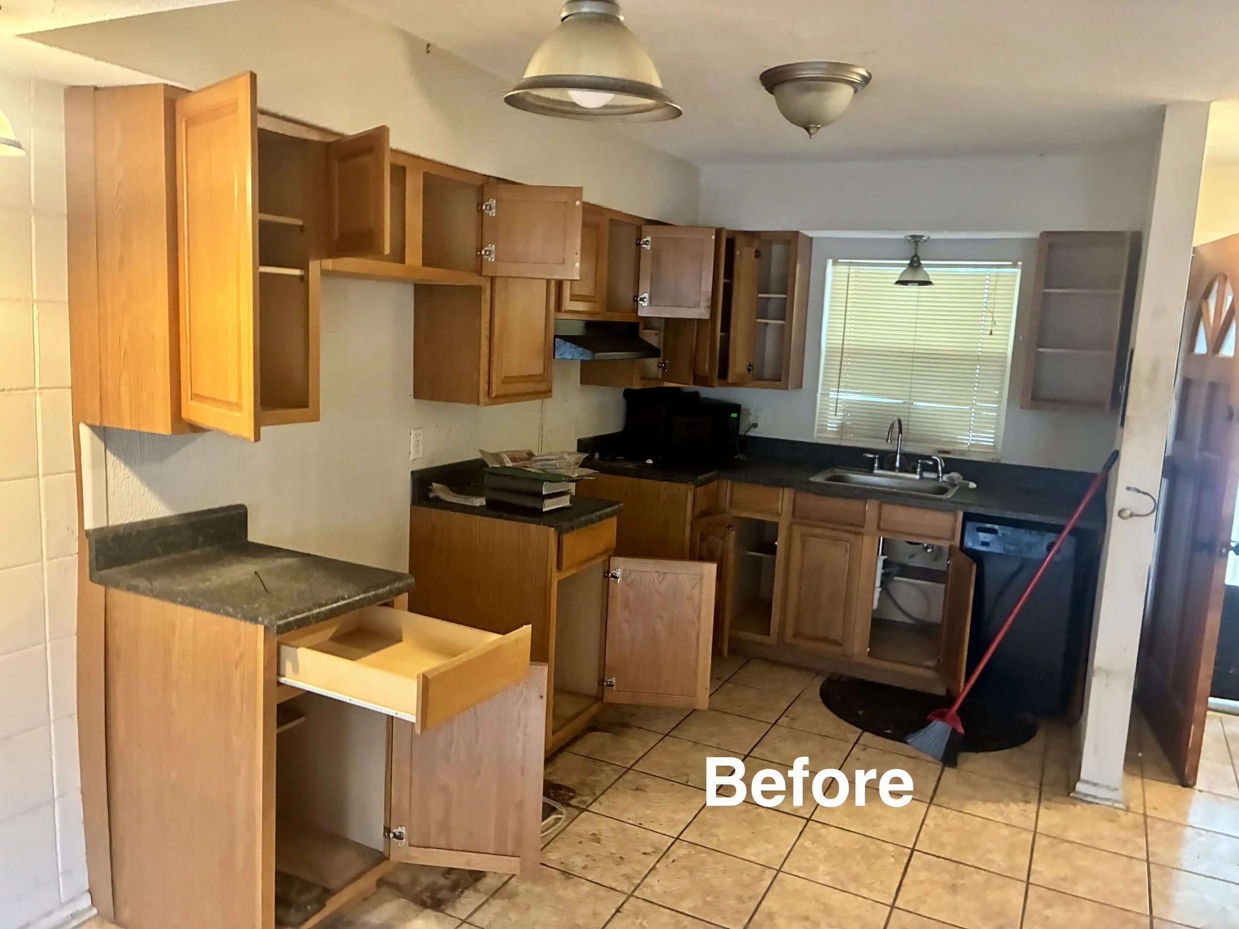 Kitchen with wooden cabinets, some open and some missing doors, a microwave on the counter, a window with blinds, and a mop leaning against the counter. The floor is tiled in a pattern.
