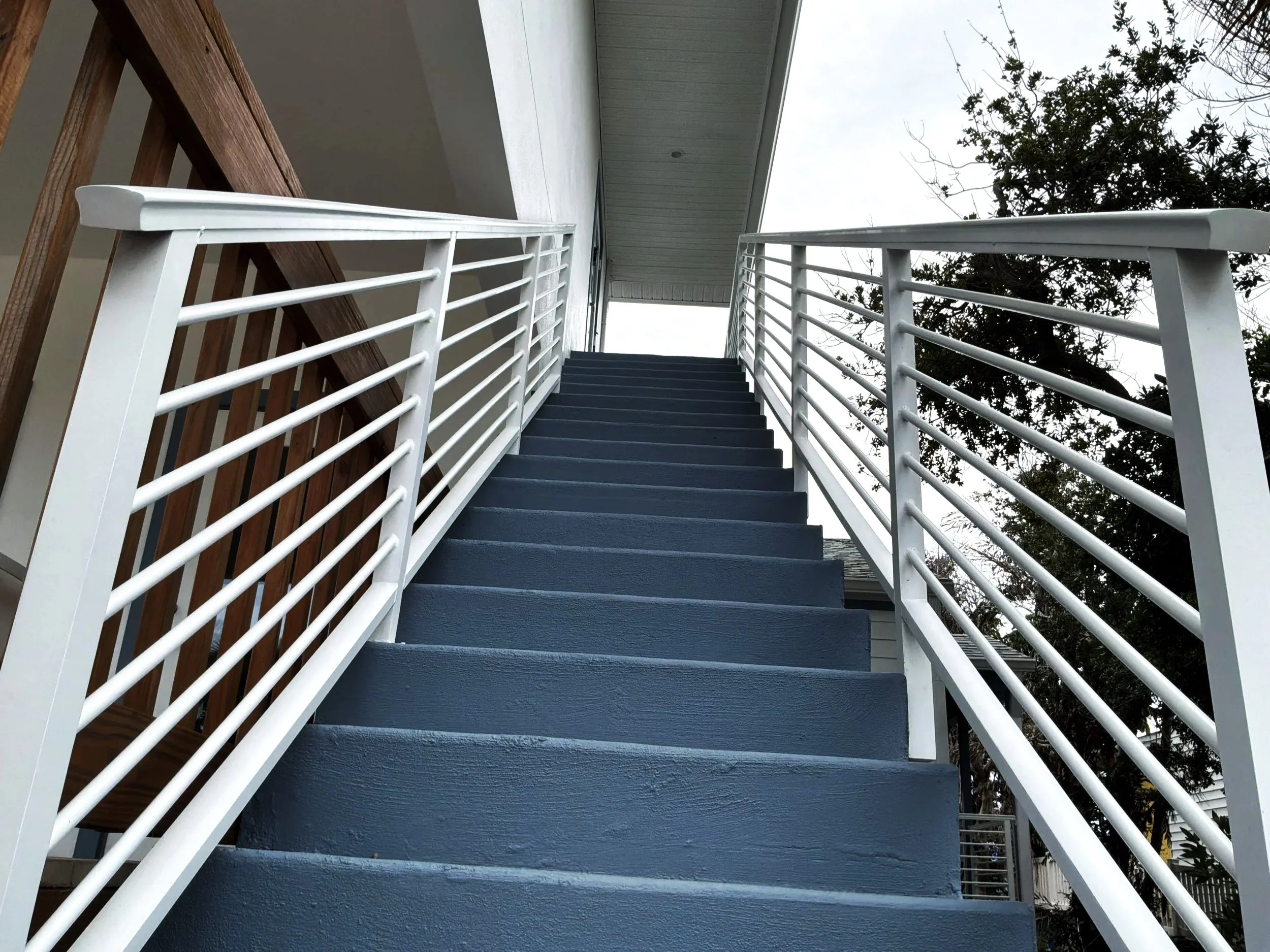 Outdoor staircase with dark blue steps, white railings, wooden side panels, and a gray ceiling, with a tree and cloudy sky in the background.