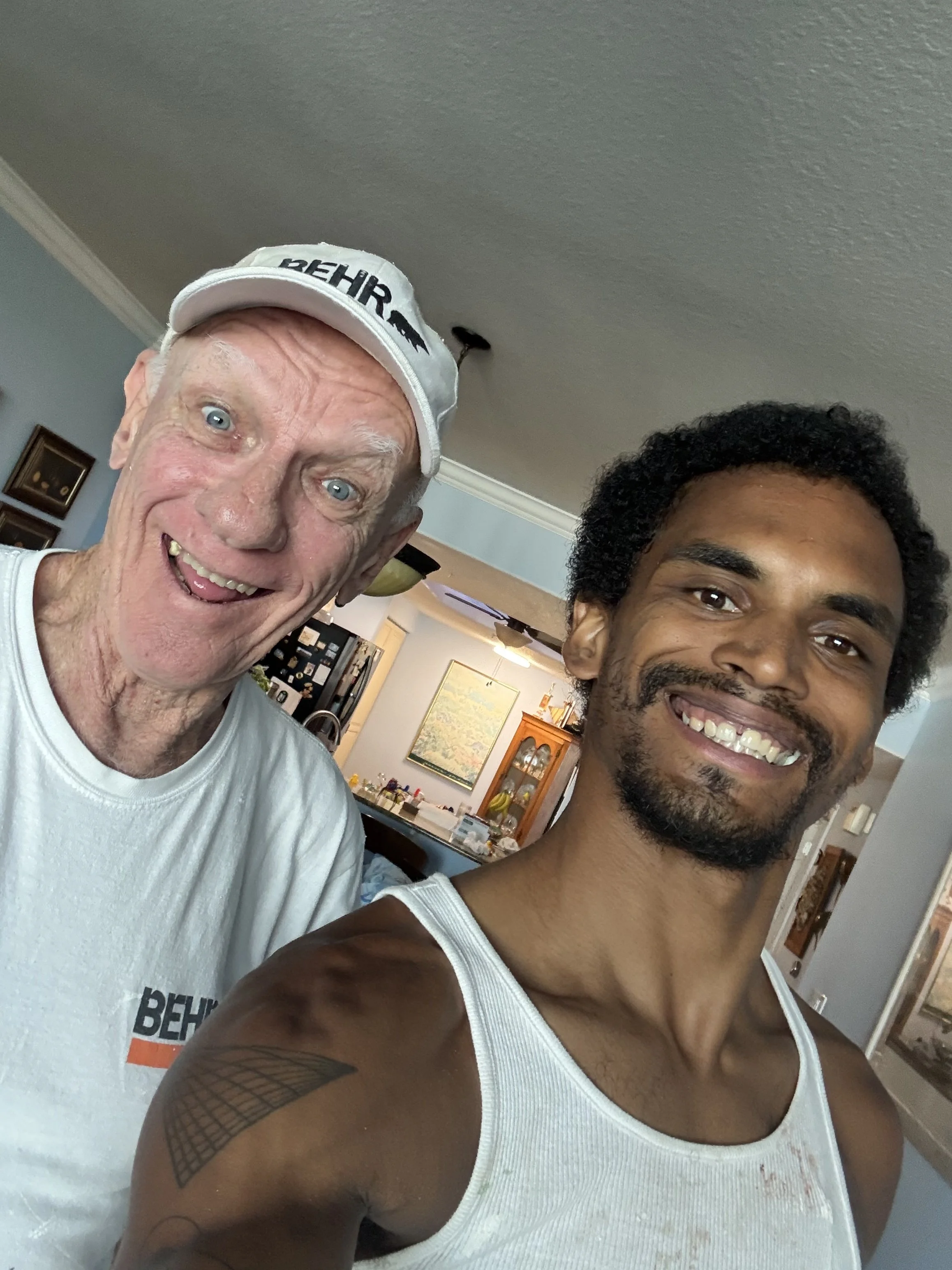 Two smiling men taking a selfie indoors, one elderly with white hair, wearing a white cap and white shirt, the other younger with curly black hair and a beard, wearing a white tank top. The background shows a kitchen with framed pictures, a map, and various kitchen items.