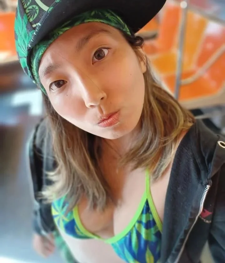 A woman taking a selfie, wearing a black cap with green palm tree design, a black jacket, and a bright blue and green swimsuit, with an orange background.