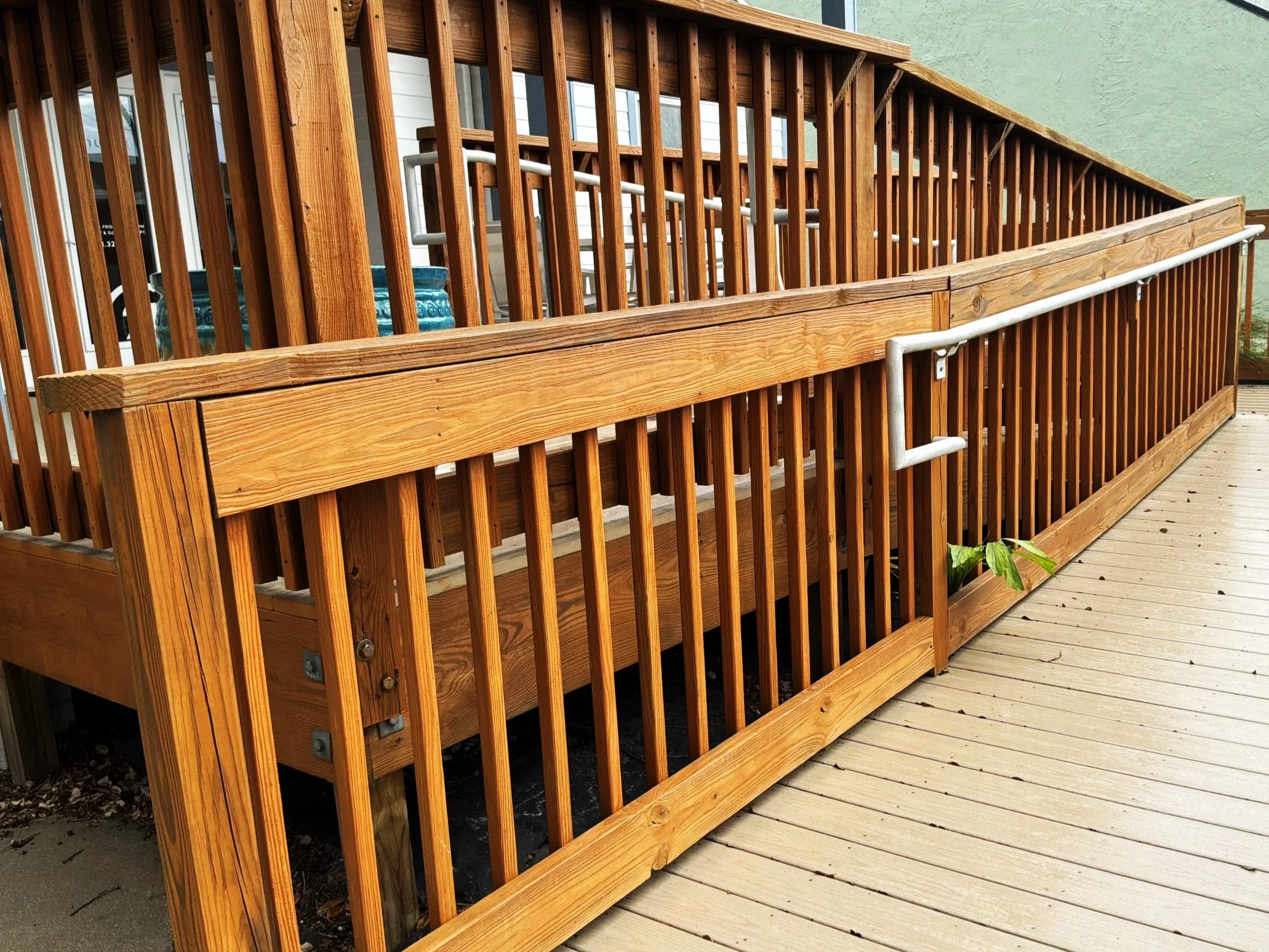 Wooden deck railing with horizontal handrail, vertical balusters, and metal brackets, next to a wooden deck floor with a small plant at the bottom edge.
