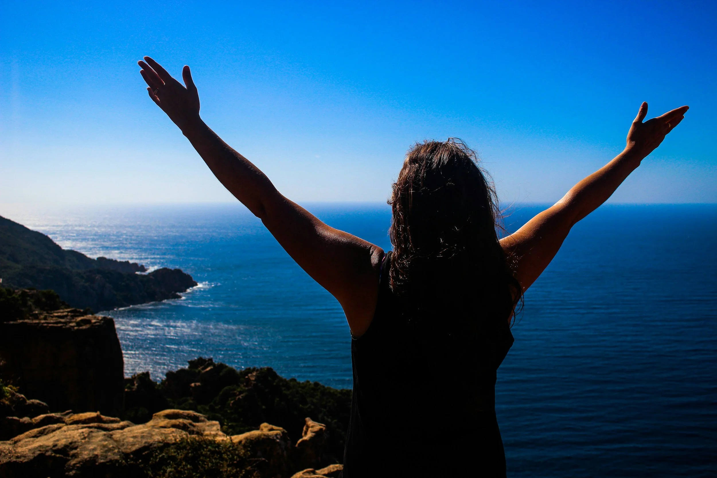 Person with long hair standing outdoors at a high vantage point with arms raised, overlooking a blue ocean and rocky coastline under clear sky.