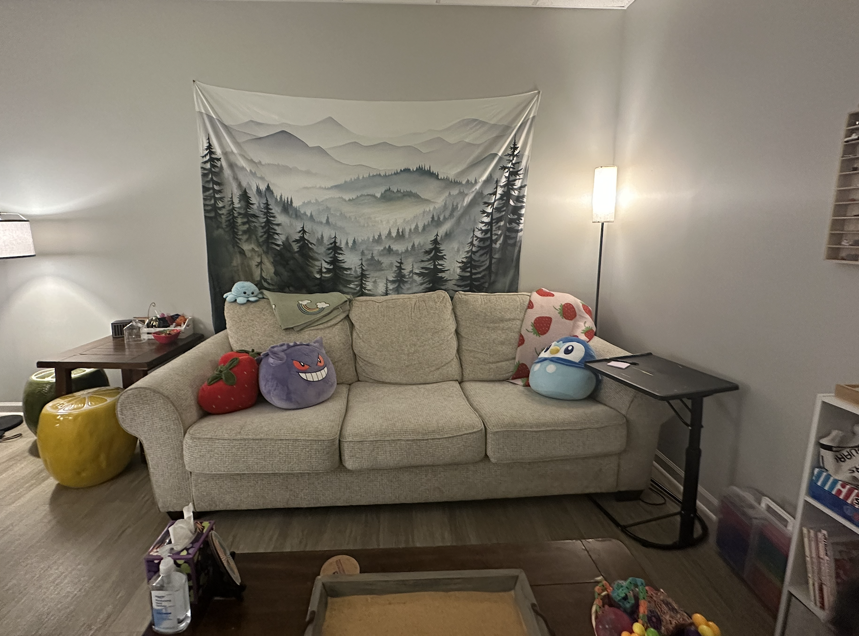 Living room with a beige couch decorated with plush toys resembling Pokémon characters, a tapestry of mountains and trees on the wall behind Additional items include a green apple-shaped ottoman, and various toys and decorations.