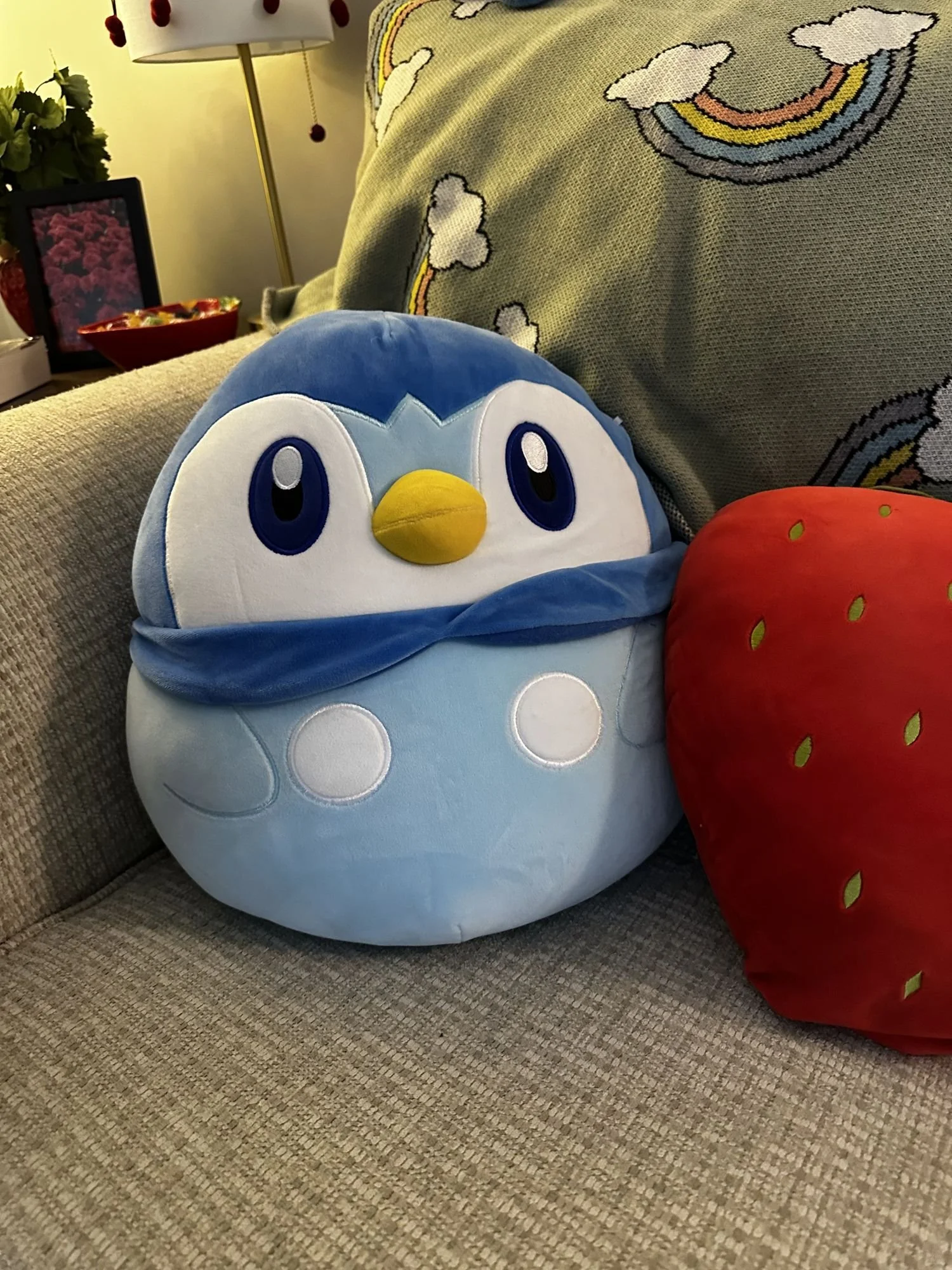 A plush stuffed penguin with blue and white coloring, large eyes, and a yellow beak, sitting on a beige textured couch.