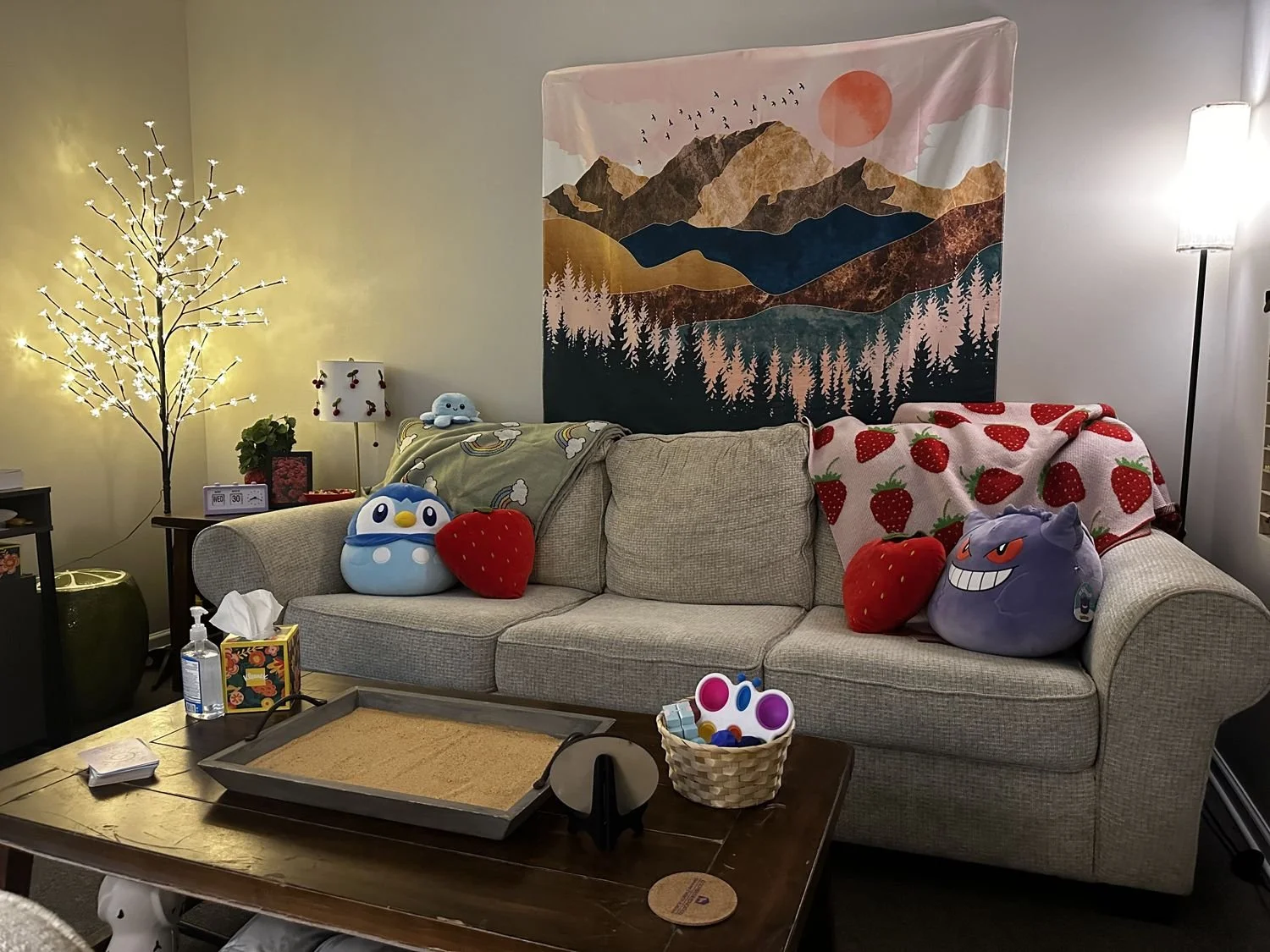 A living room with a beige sofa decorated with strawberry-shaped plush pillows and character pillows, a tapestry featuring mountains and a sunset on the wall, a small lamp with cherry decoration, and a tree-shaped light with small white lights. A tab