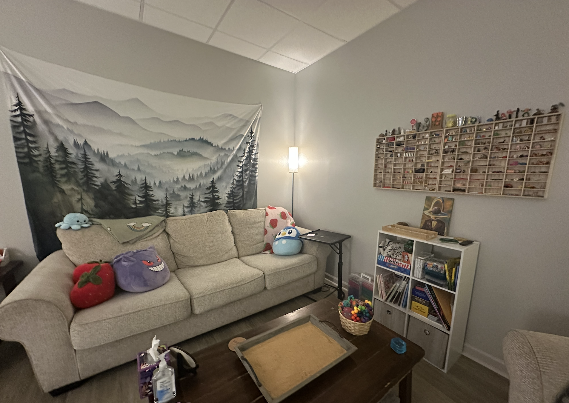 A living room with a beige couch decorated with plush Pokémon toysThere is a tapestry of a misty mountain landscape hanging on the wall, a tall floor lamp, and a wall-mounted wooden shelf filled with small collectibles. A small white storage cube hol